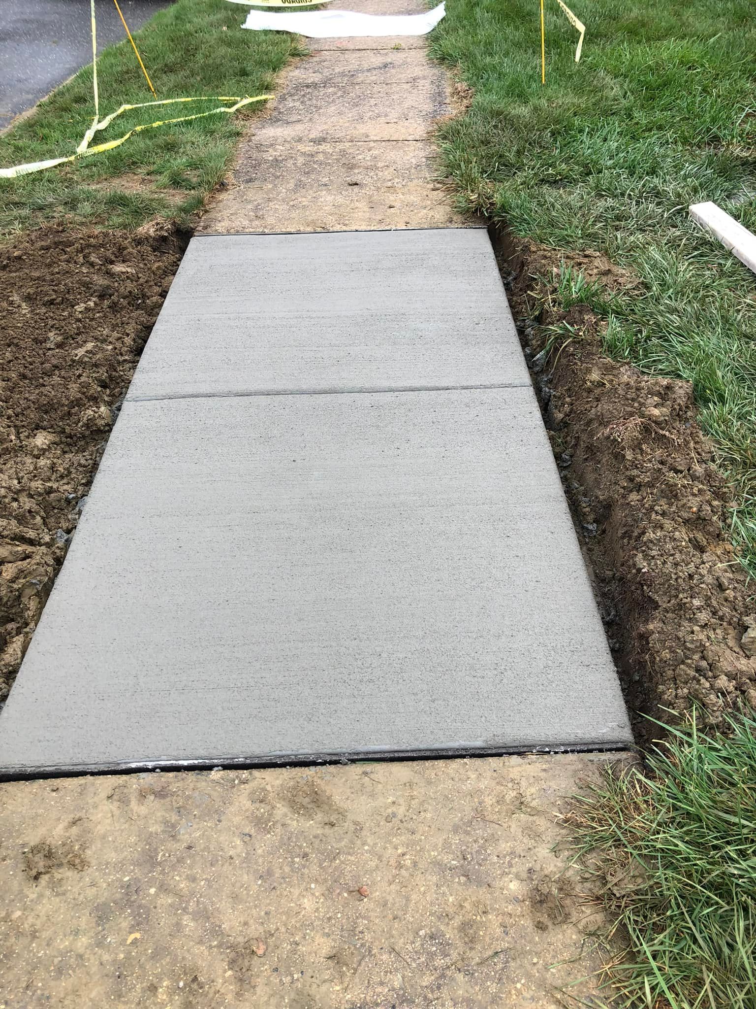 A newly poured section of smooth gray concrete sidewalk centered between older, weathered pavement and grassy edges.