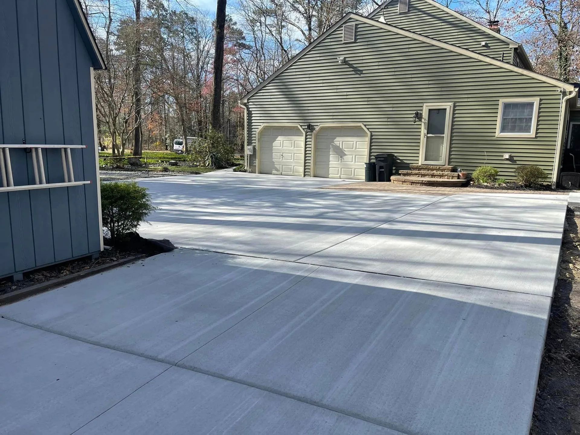 A newly poured, light gray concrete driveway leads up to a garage with two doors and a house with green siding.