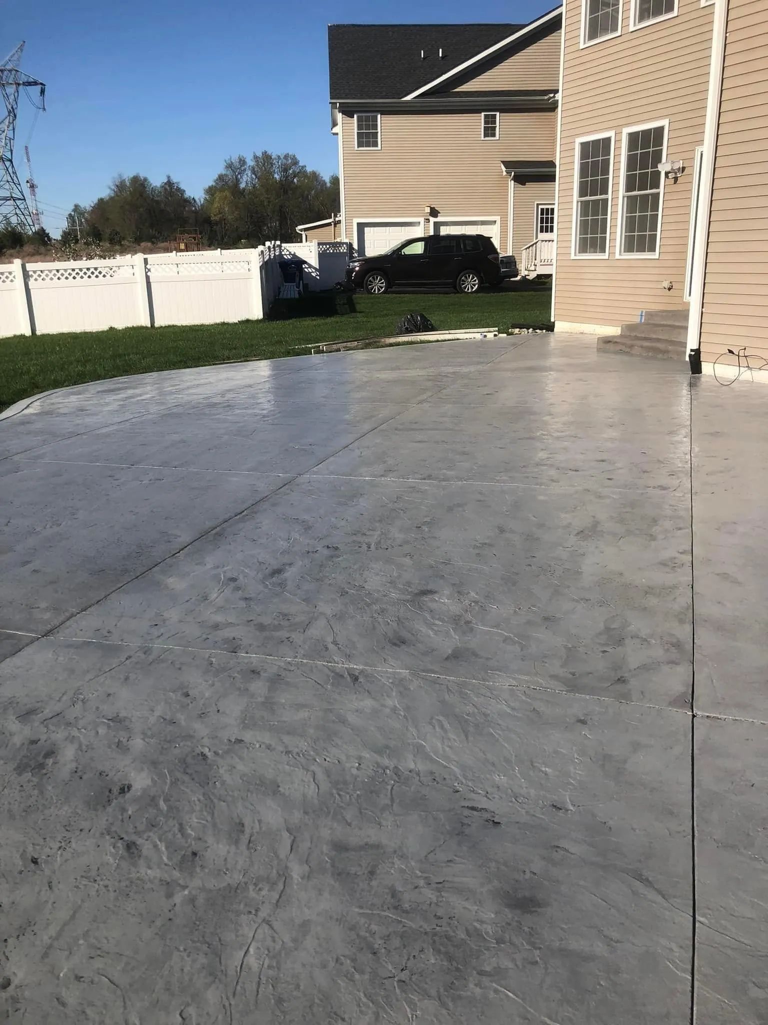 A gray, patterned concrete patio in a backyard with a house, white fence, and lawn in the background.