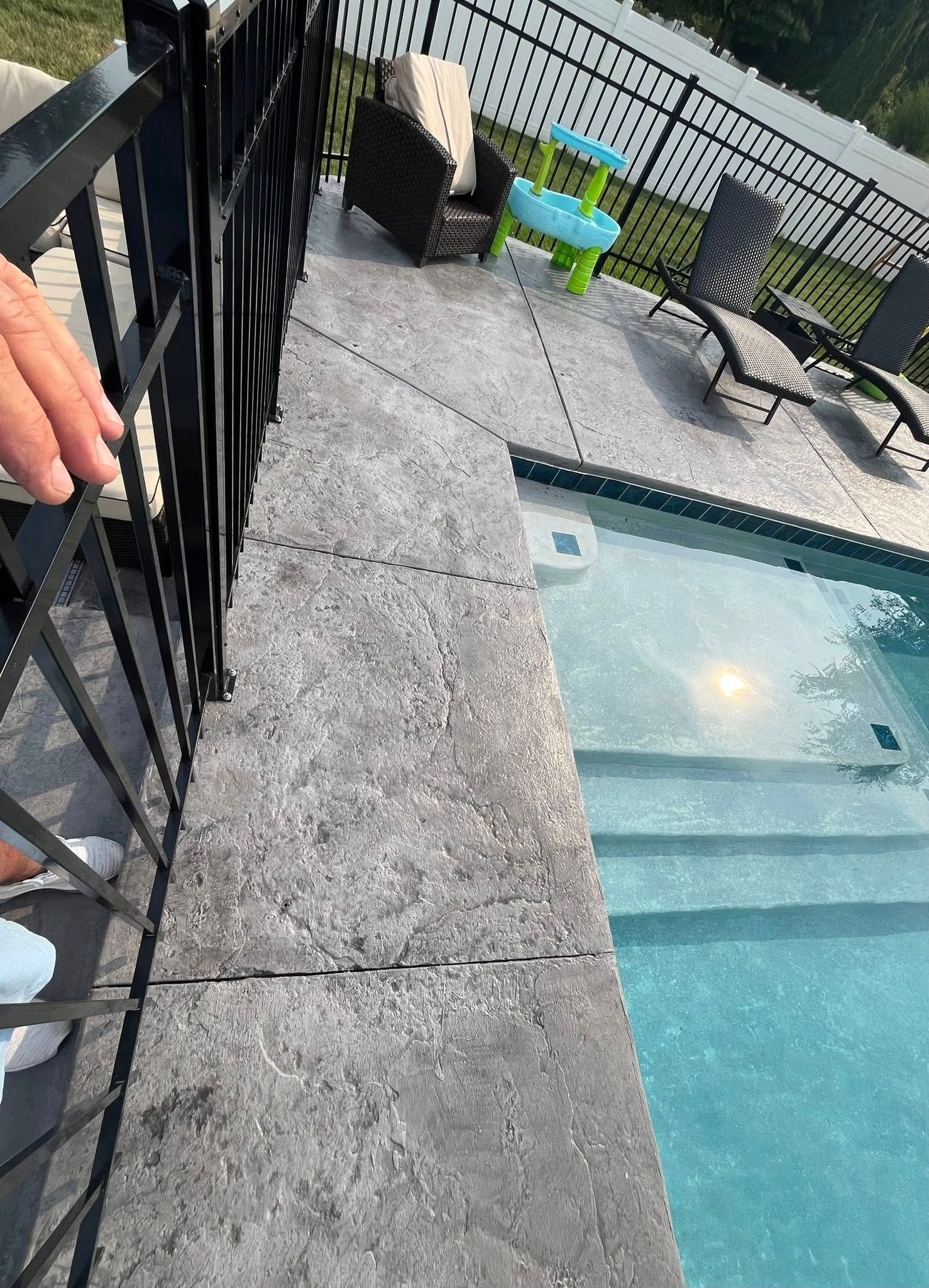 A person's hand rests on a black pool fence overlooking a pool deck with lounge chairs and a small pool slide.