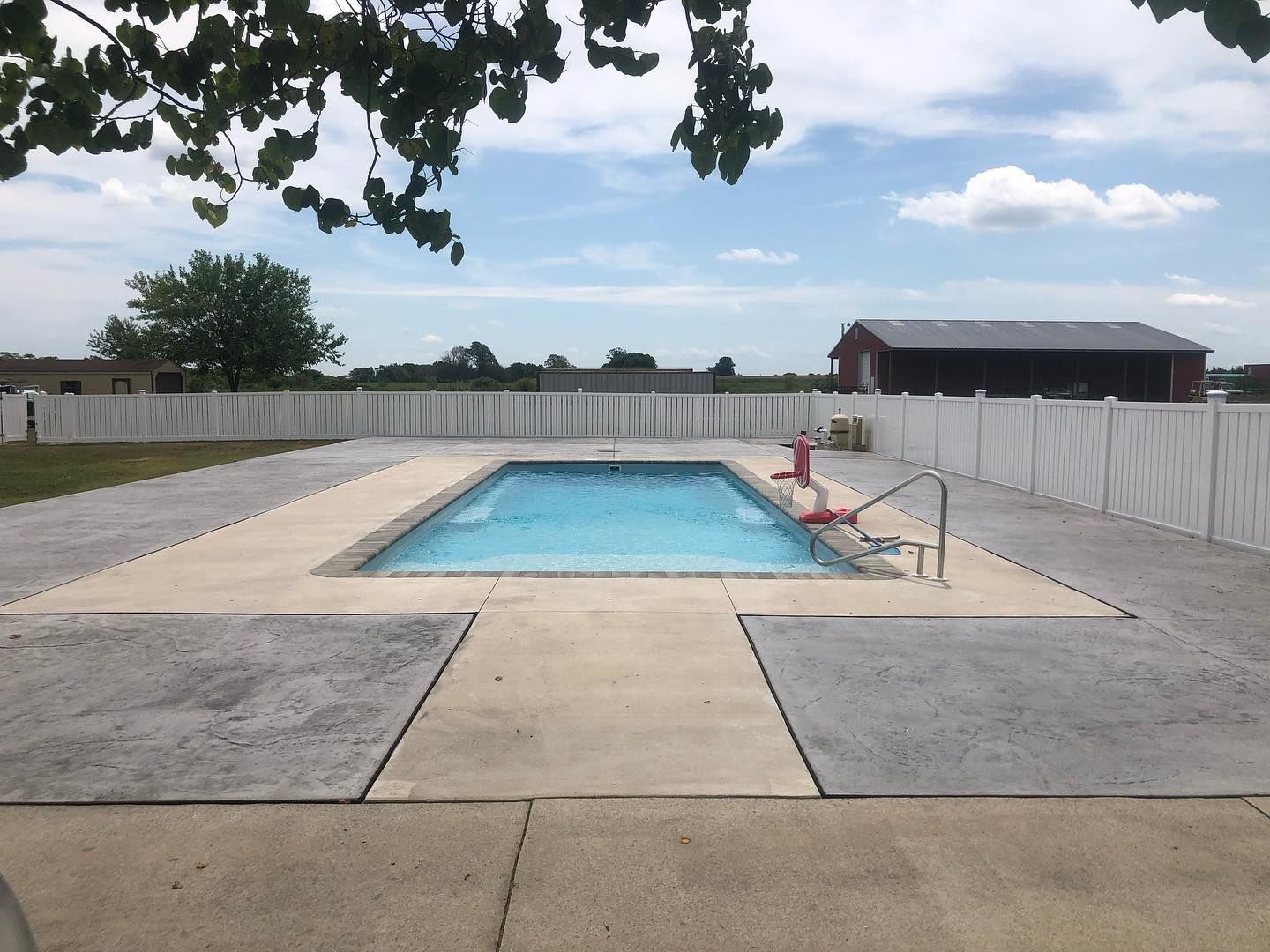 A rectangular swimming pool surrounded by a light-colored concrete deck, enclosed by a white fence under a cloudy sky.