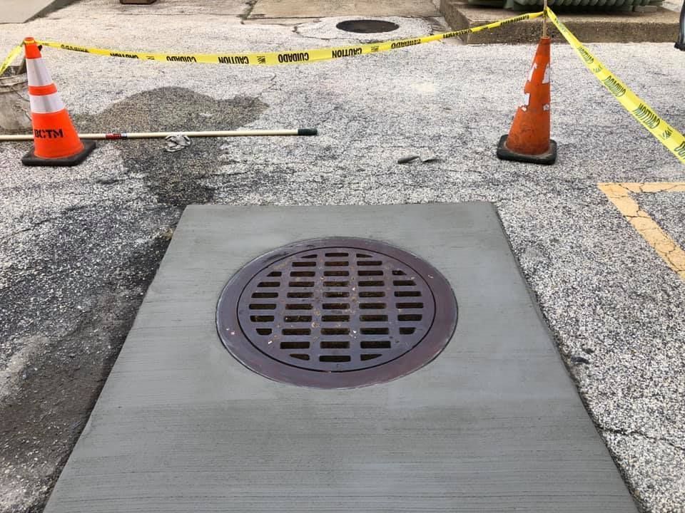 A new concrete square patch surrounds a circular metal storm drain grate, marked by two traffic cones and caution tape.