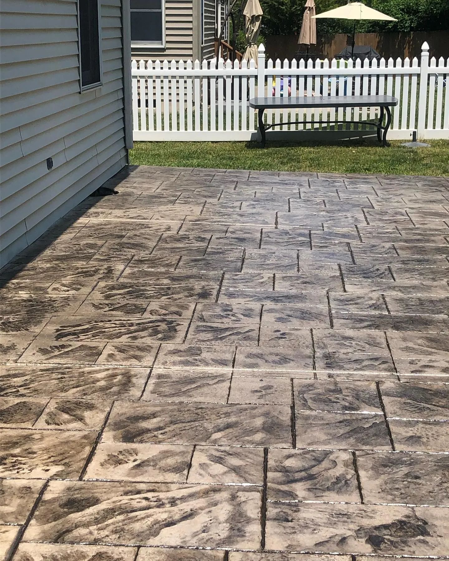 A patterned, brown and gray stone-look concrete patio next to a house, with a white picket fence in the background.