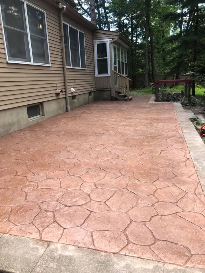 A tan house with a stamped, reddish-brown concrete patio featuring a flagstone pattern, bordering a wooded backyard.