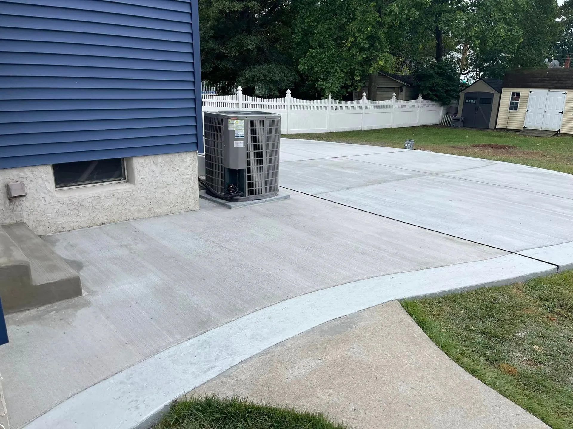 A freshly poured concrete patio adjacent to a blue house with an air conditioning unit and a white fence in the backyard.