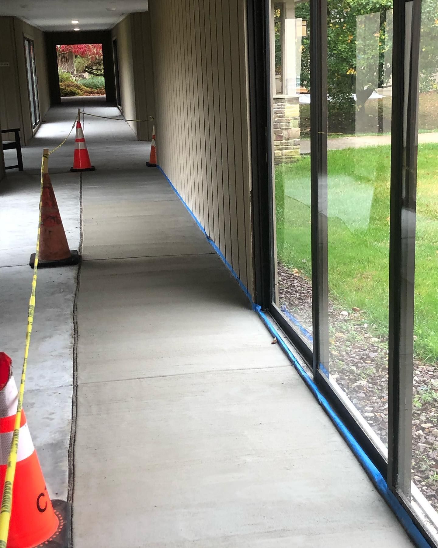 A narrow concrete walkway under construction next to a glass wall, with orange safety cones blocking off the area.