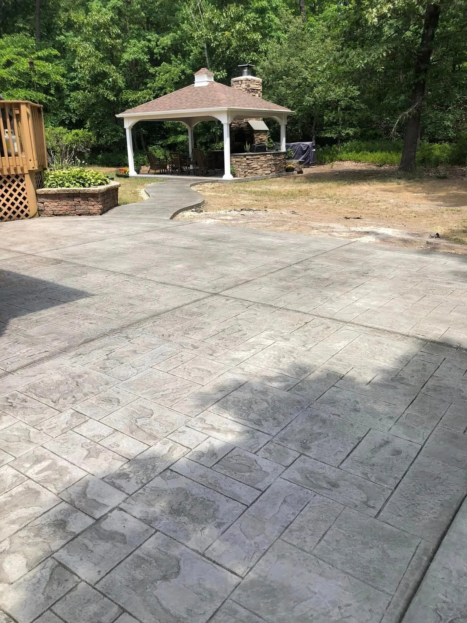 A backyard patio with gray stamped concrete pavers leading to a white gazebo with a stone fireplace, surrounded by trees.