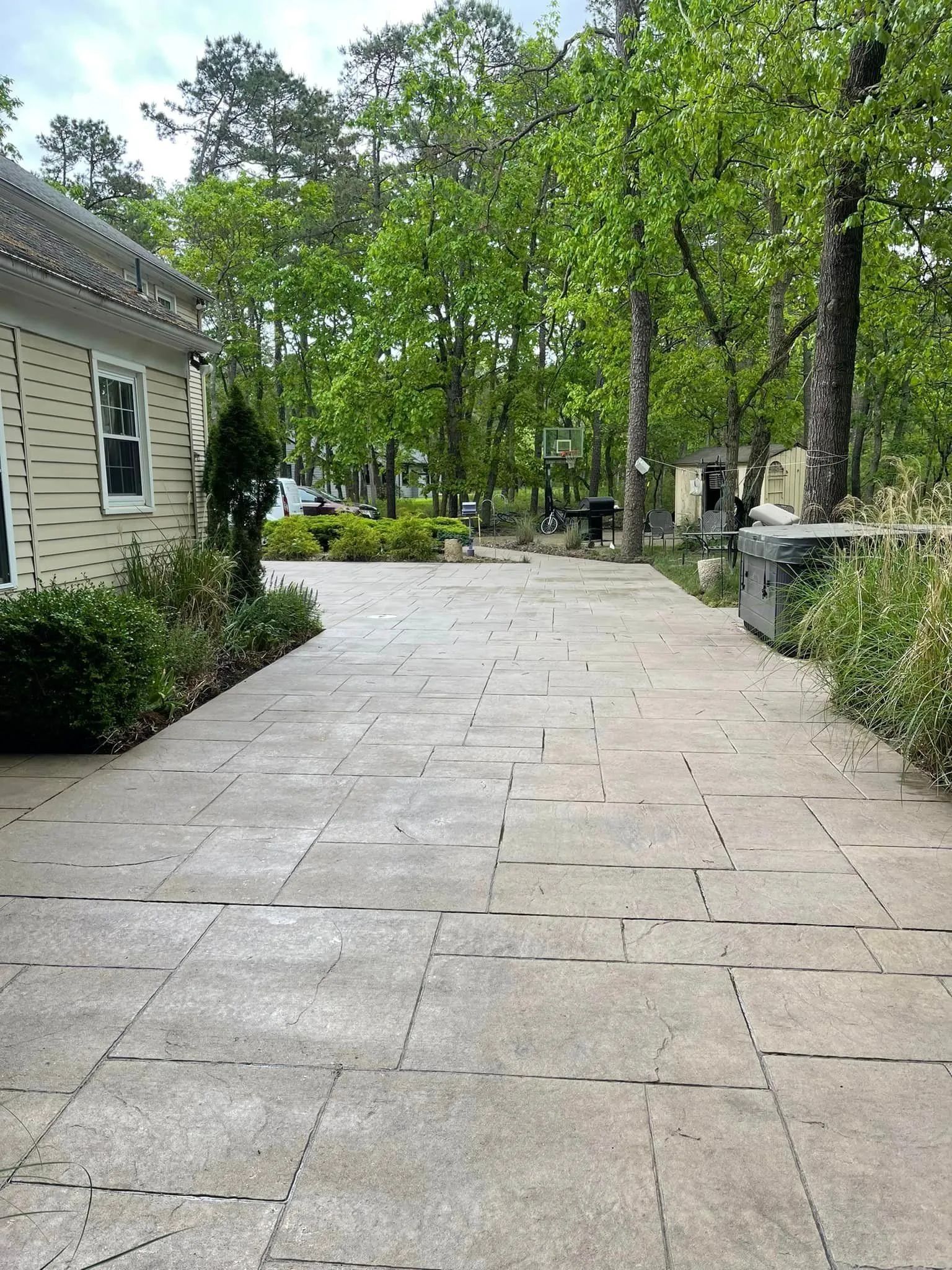 A light gray concrete patio with dark square accent inlays, next to a beige building, leading toward a wooded backyard.