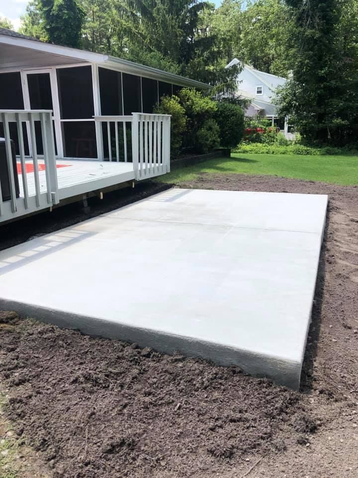 A newly poured, rectangular concrete patio sits on bare dirt next to a white deck and screened-in porch.