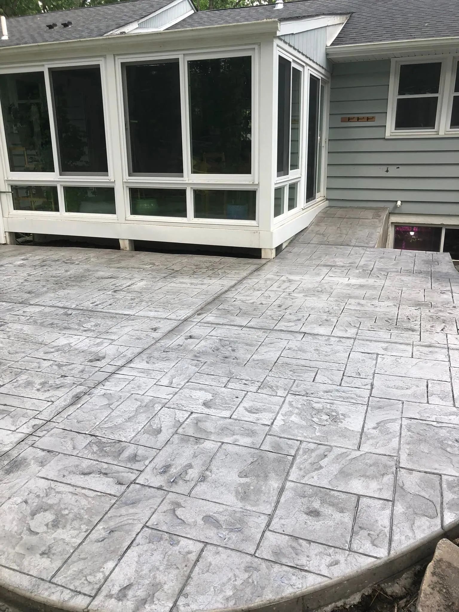 A gray-toned stamped concrete patio with a rectangular block pattern, adjacent to a house with a white-framed sunroom.