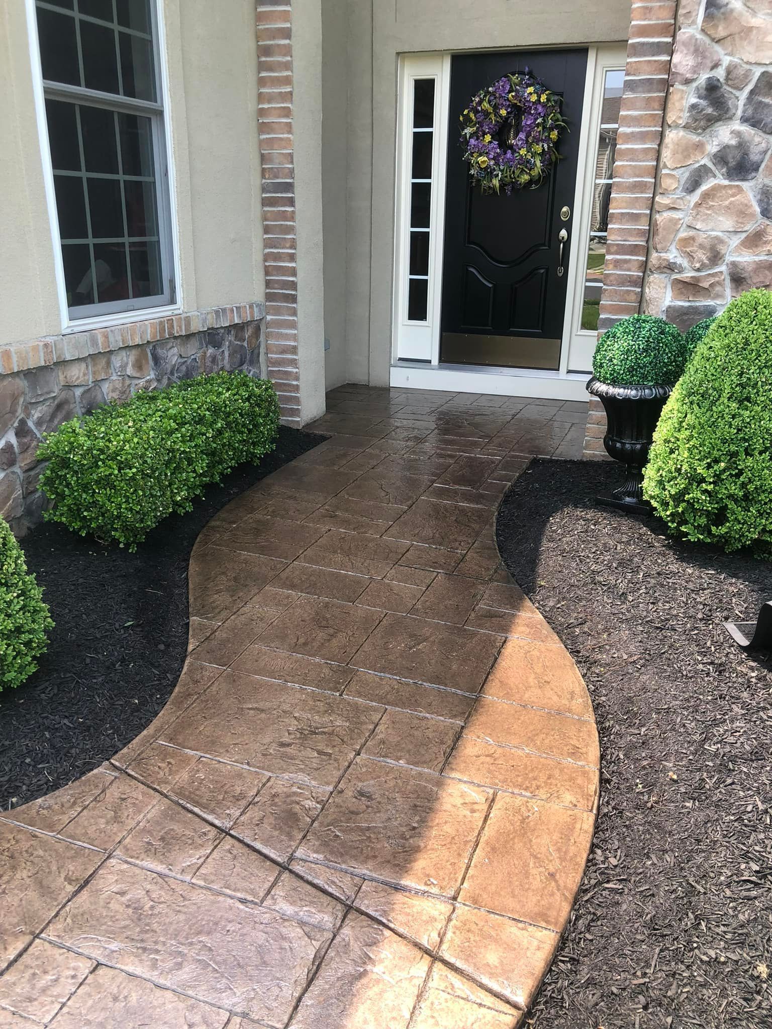 A stone walkway leads to a front door with a wreath, flanked by manicured green shrubs and dark landscaping mulch.