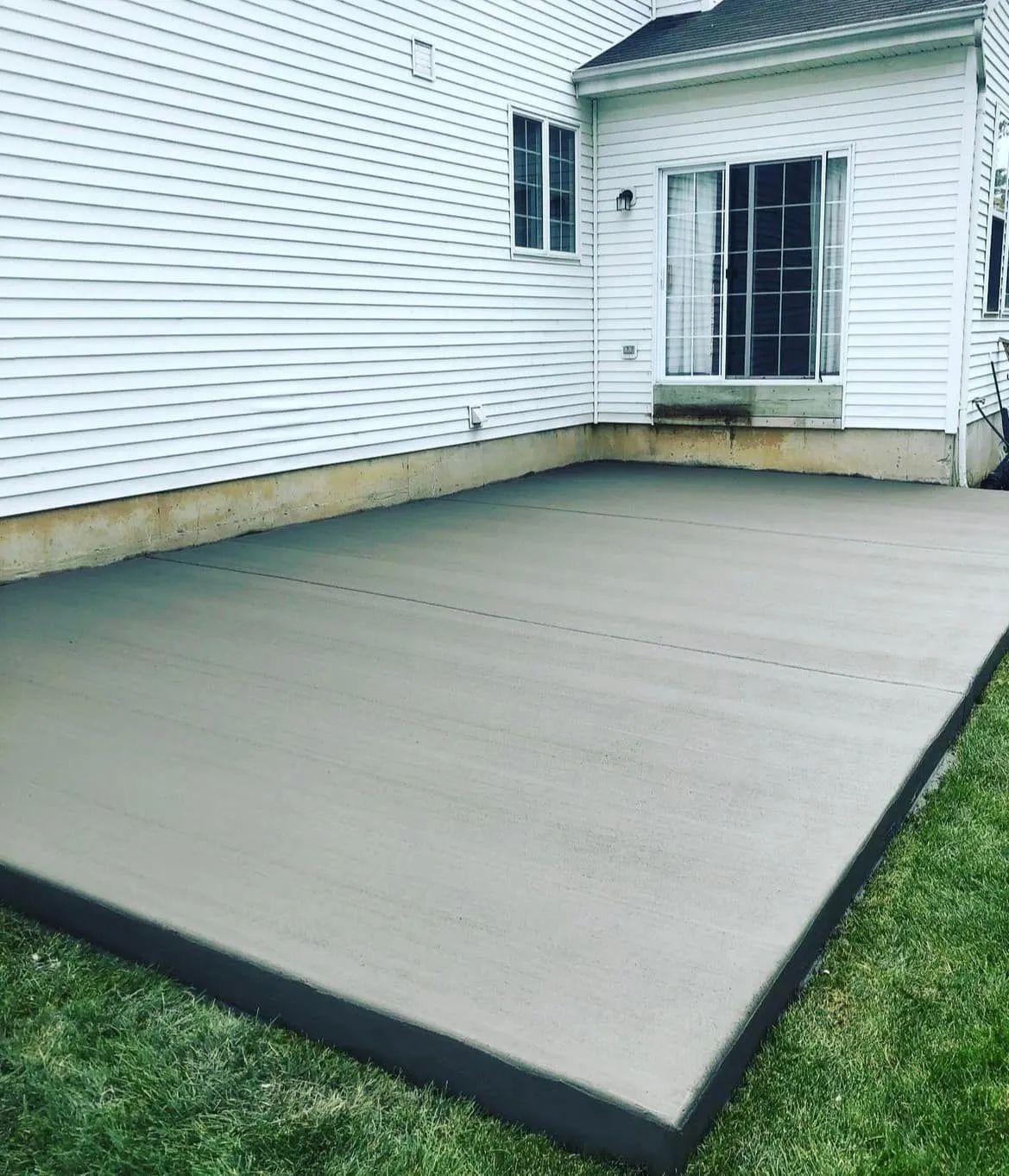 A new rectangular concrete patio installed against the side of a white-sided house with a sliding glass door.