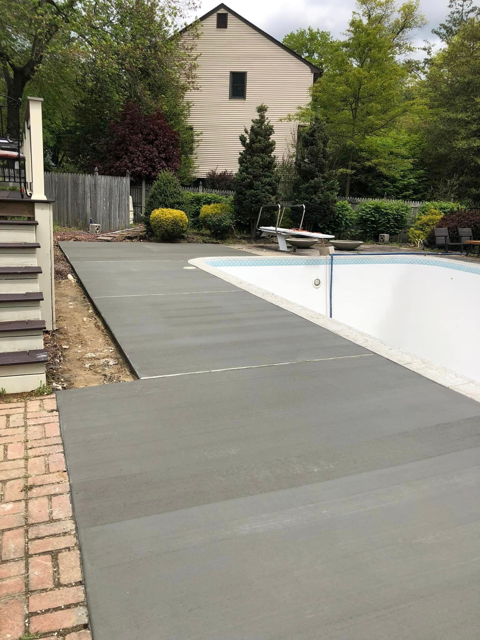 A newly poured gray concrete pool deck walkway stretching alongside an empty swimming pool in a residential backyard.