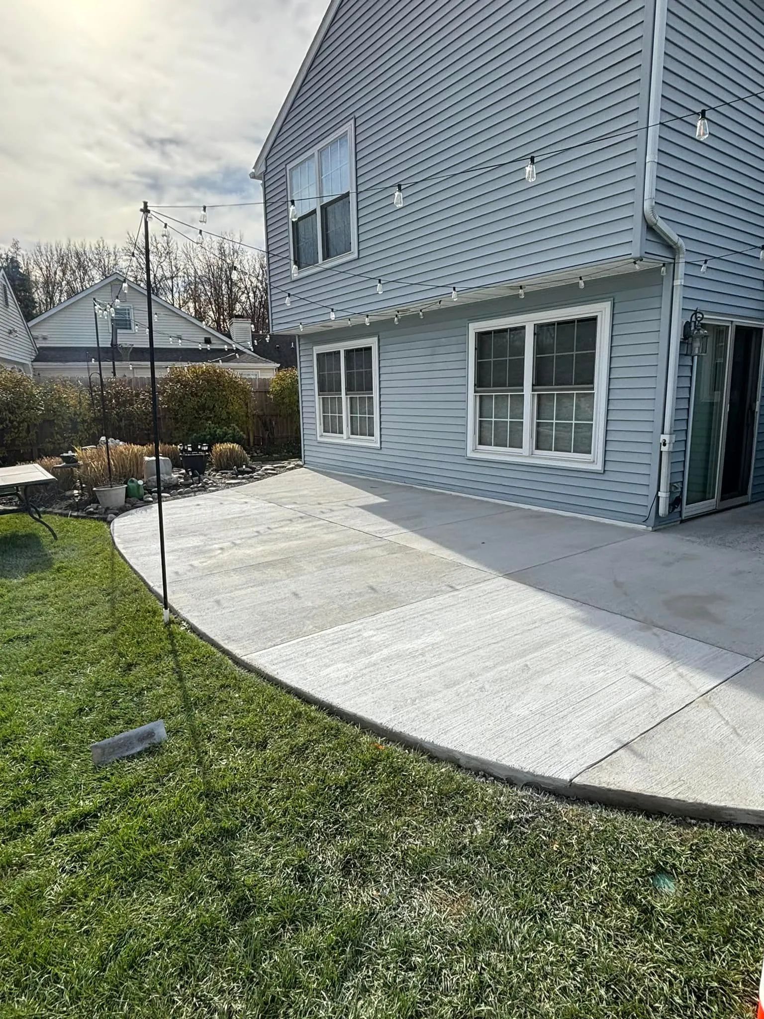 Light blue siding house with a curved concrete patio and string lights in a backyard with a grassy lawn.