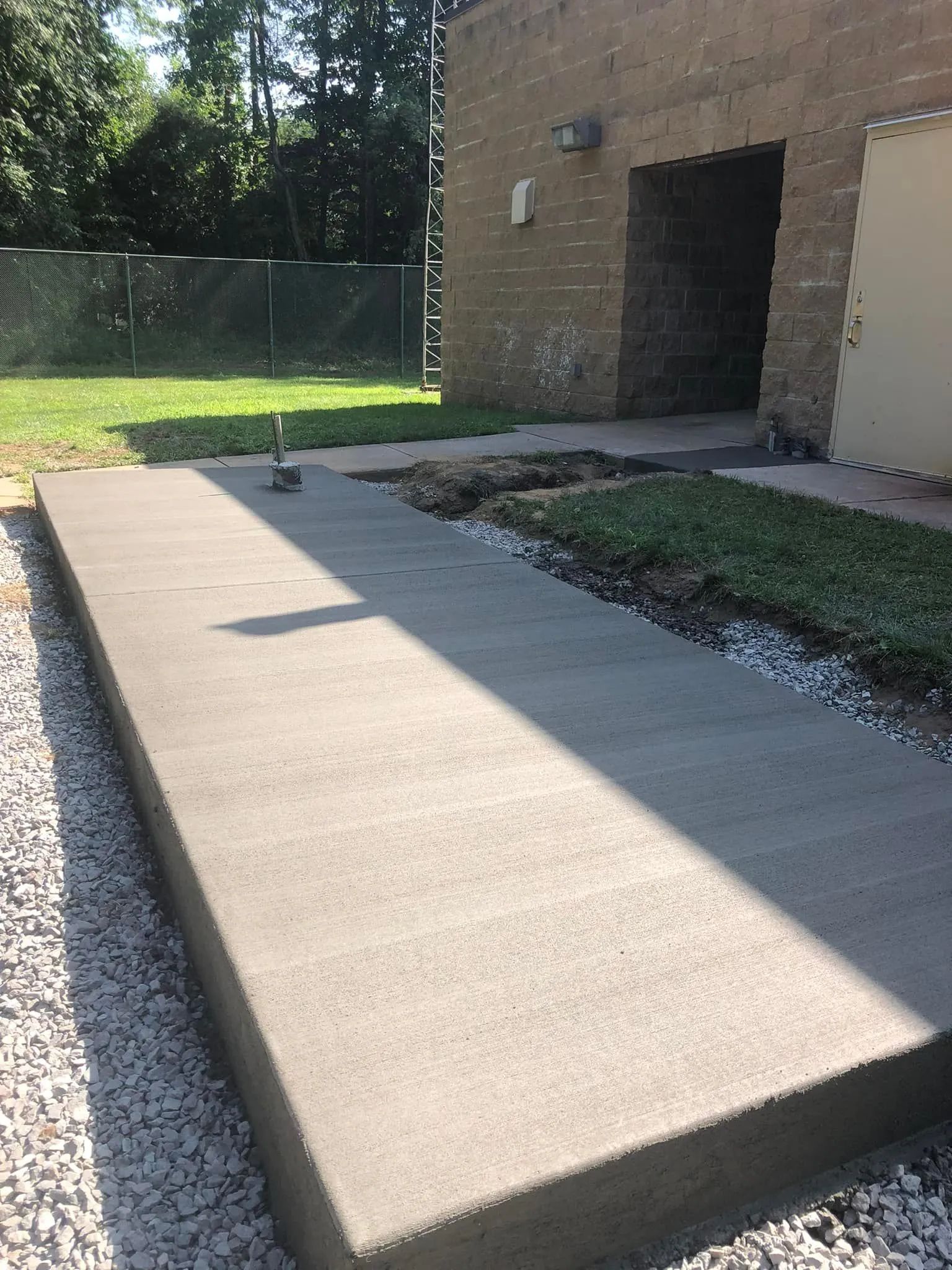 A newly poured concrete rectangular slab sits on a gravel base next to a brick building wall.