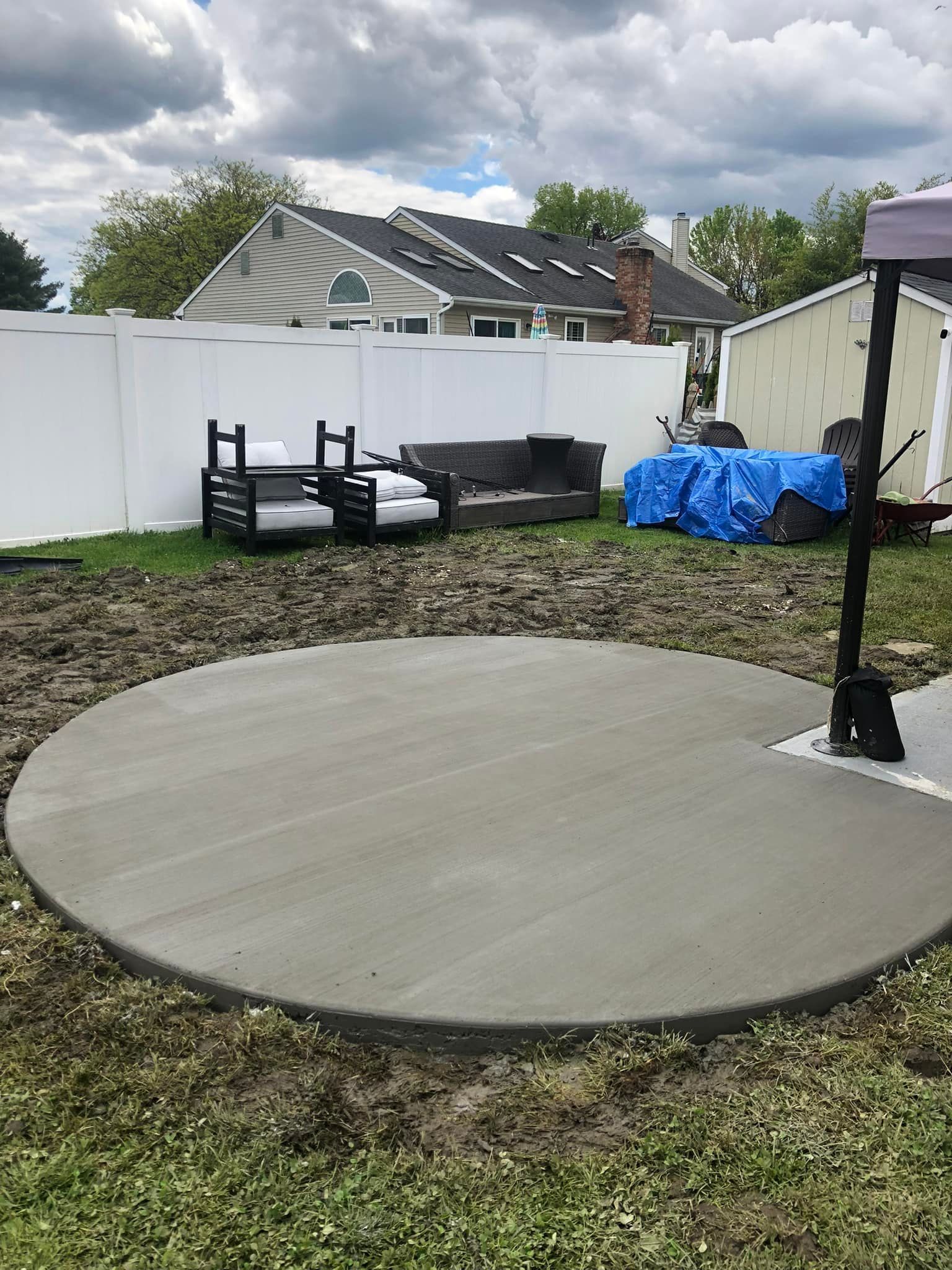 A newly poured circular concrete patio in a grassy backyard with patio furniture and a blue-covered item in the background.