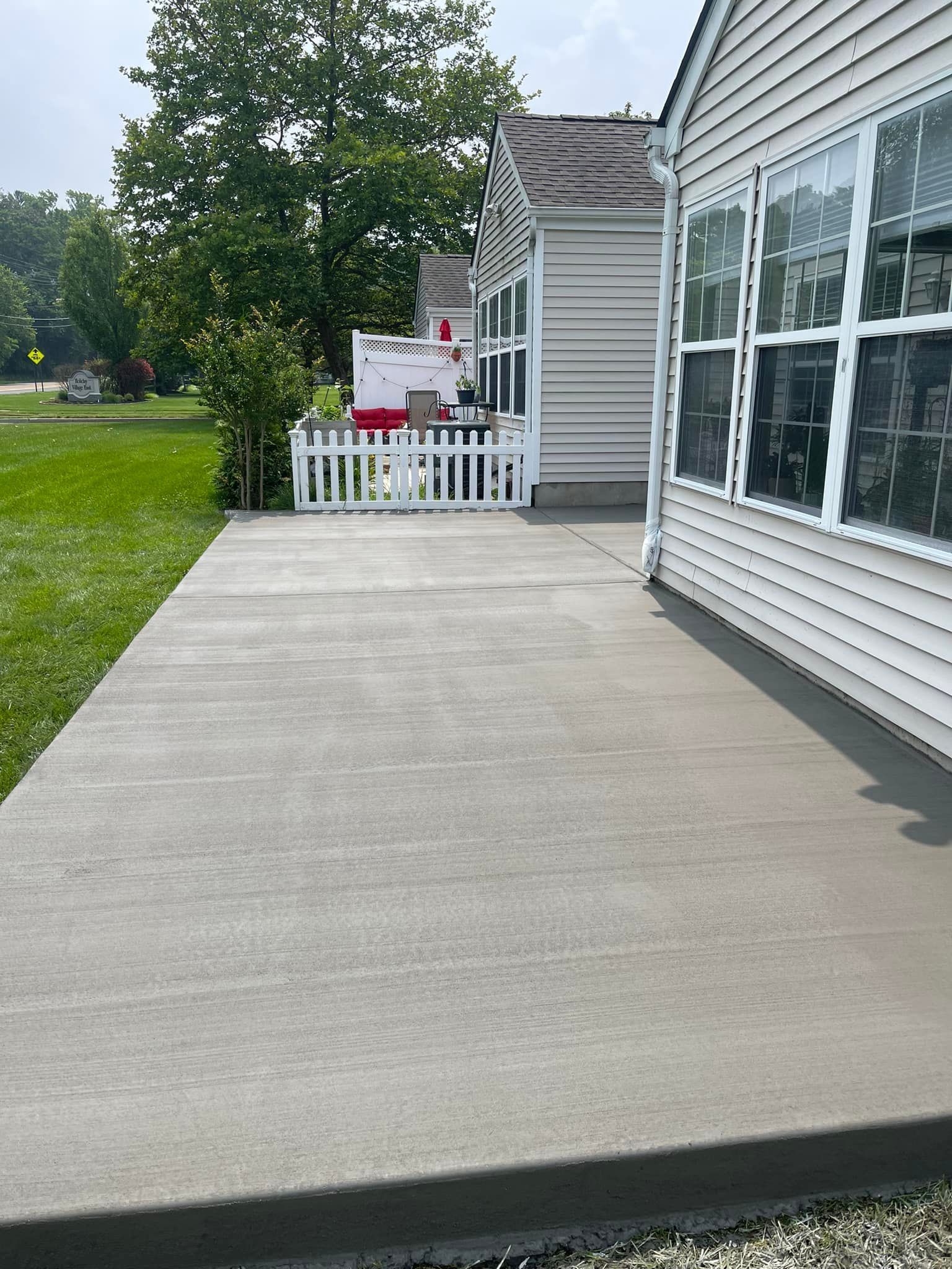 A wide, light gray concrete walkway runs alongside a white vinyl-sided house with windows and a small fenced-in porch.