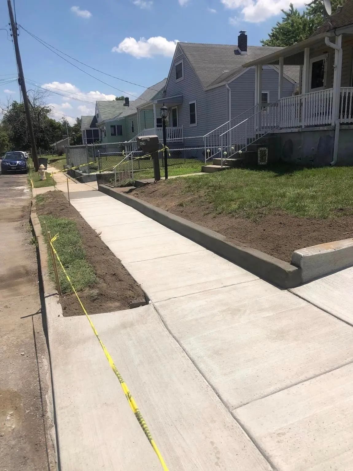A newly poured concrete sidewalk runs along a row of houses, with a yellow caution tape lining the edge of the grass.