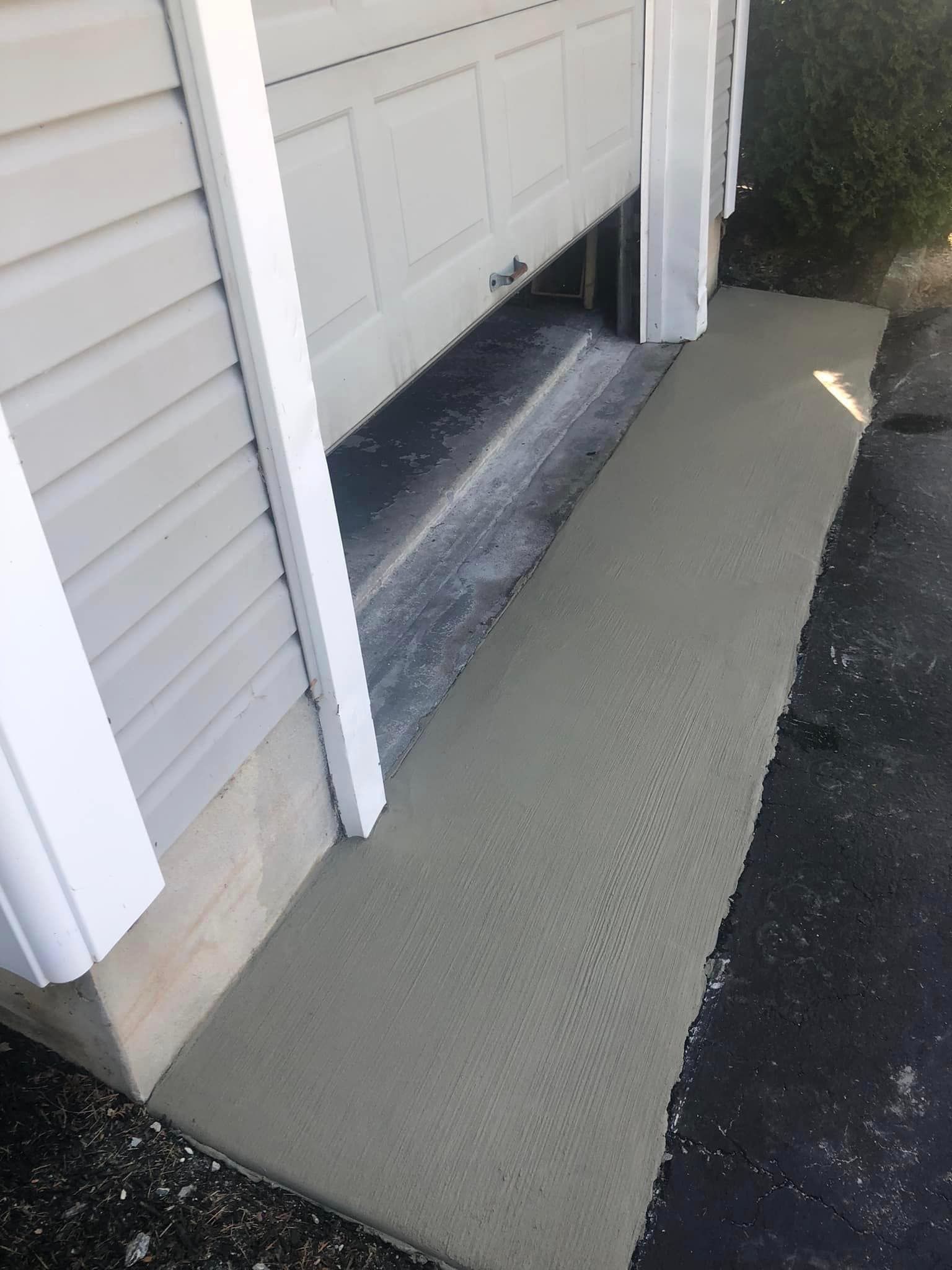 Freshly poured concrete ramp leading up to a garage door entrance next to an asphalt driveway.