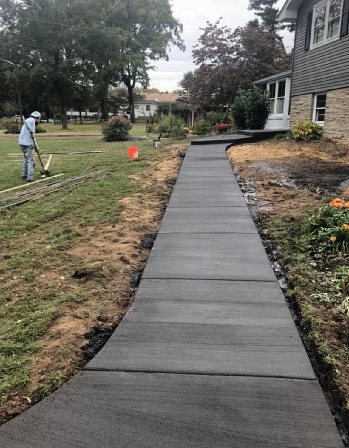 A newly poured concrete walkway leads to the side entrance of a gray house, with a worker landscaping in the yard nearby.