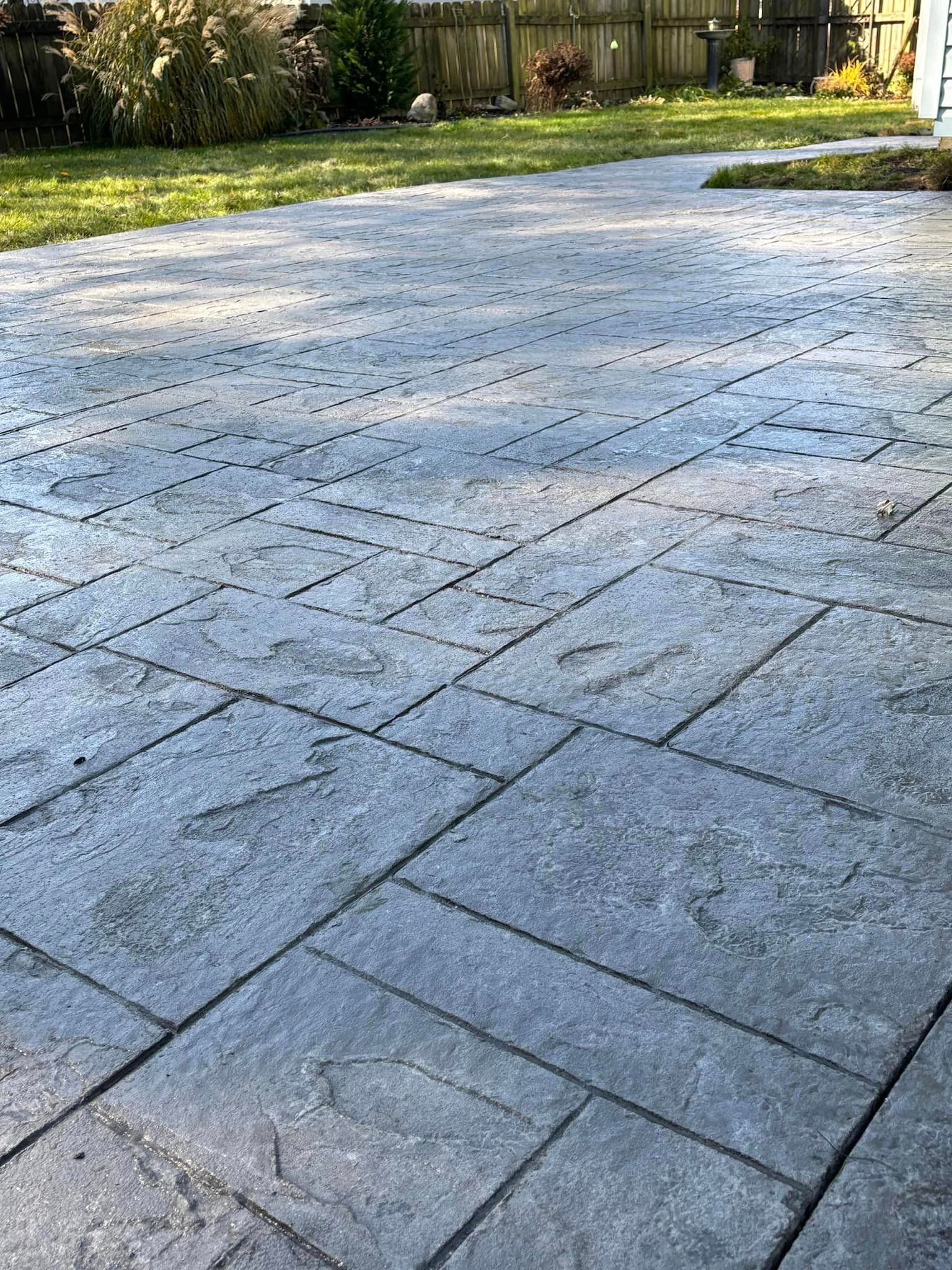 A light gray stamped concrete patio with a rectangular pattern, set against a green lawn and a wooden fence.