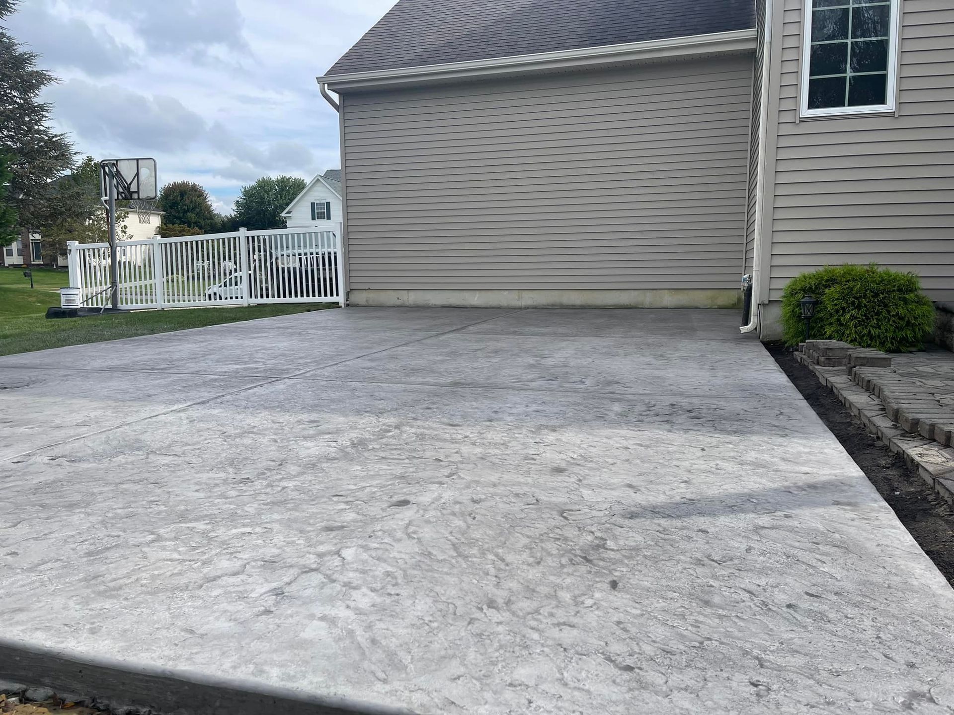 A stamped concrete patio extending from the side of a house with light gray vinyl siding and a small green bush.
