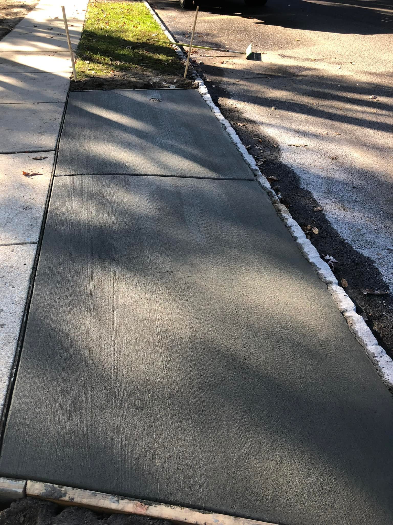 A freshly poured concrete sidewalk section between an existing older sidewalk, a patch of grass, and a dirt shoulder.