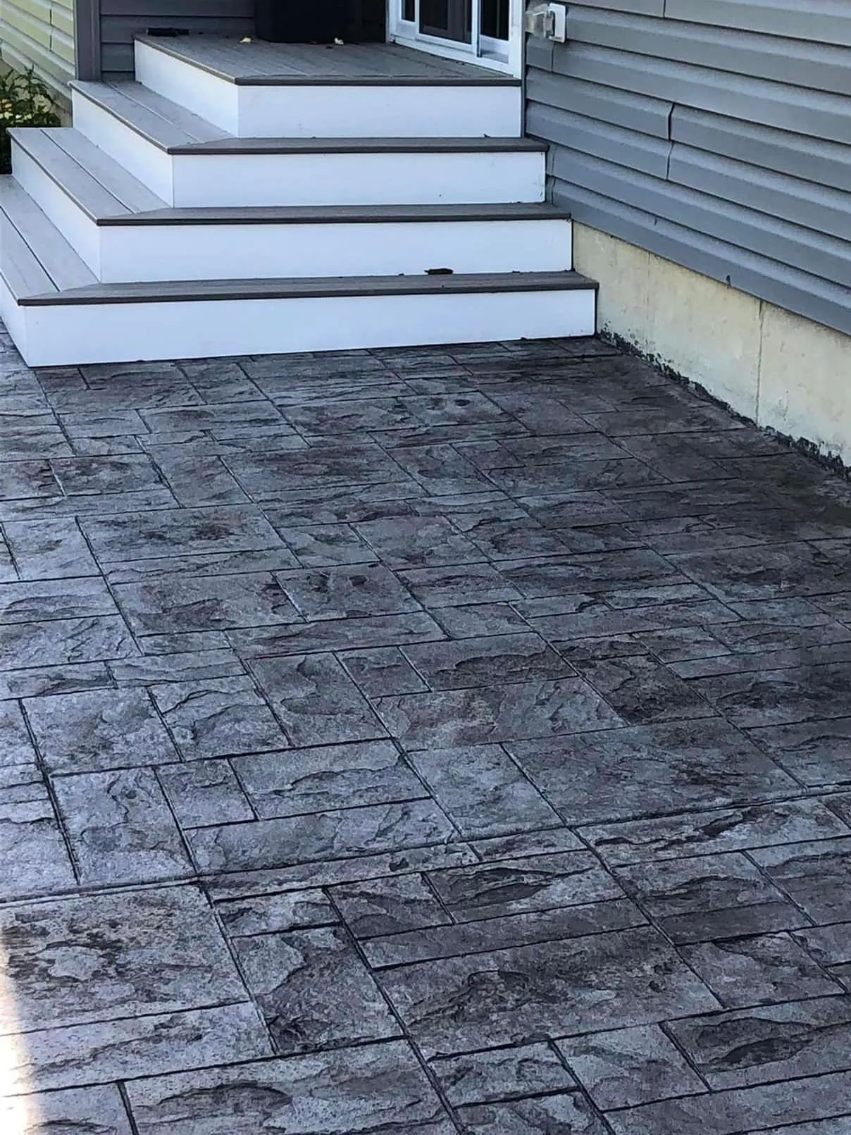 A dark, textured stamped concrete patio leading to a set of three white-trimmed, gray wooden stairs by a house exterior.