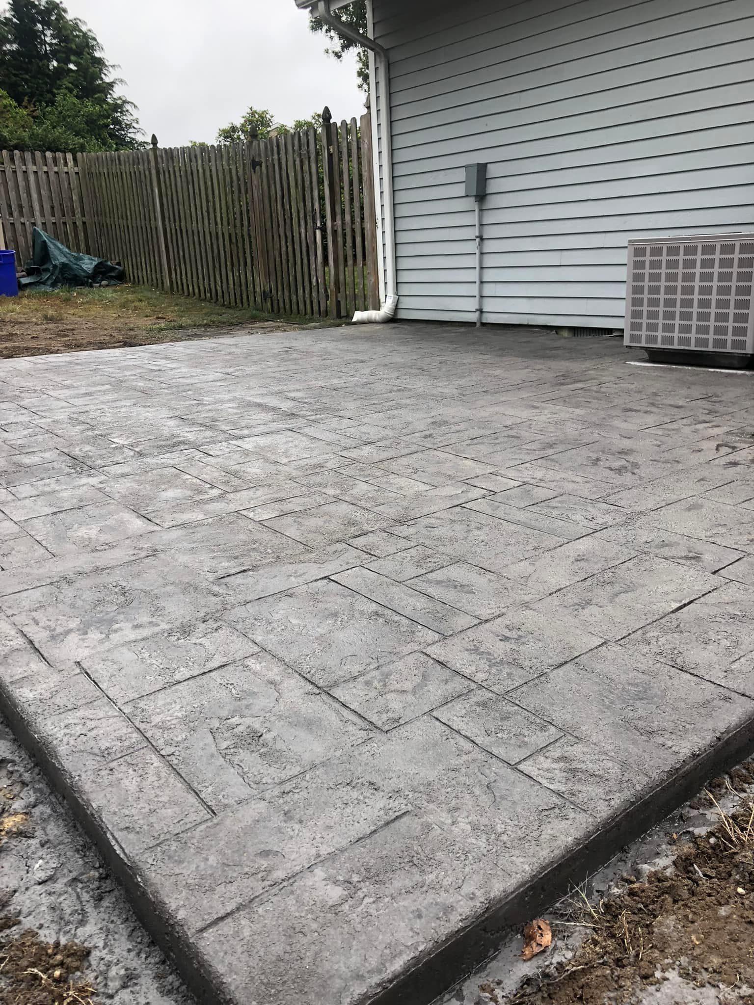 A newly poured, gray stamped concrete patio with a rectangular block pattern, adjacent to a house wall and wood fence.