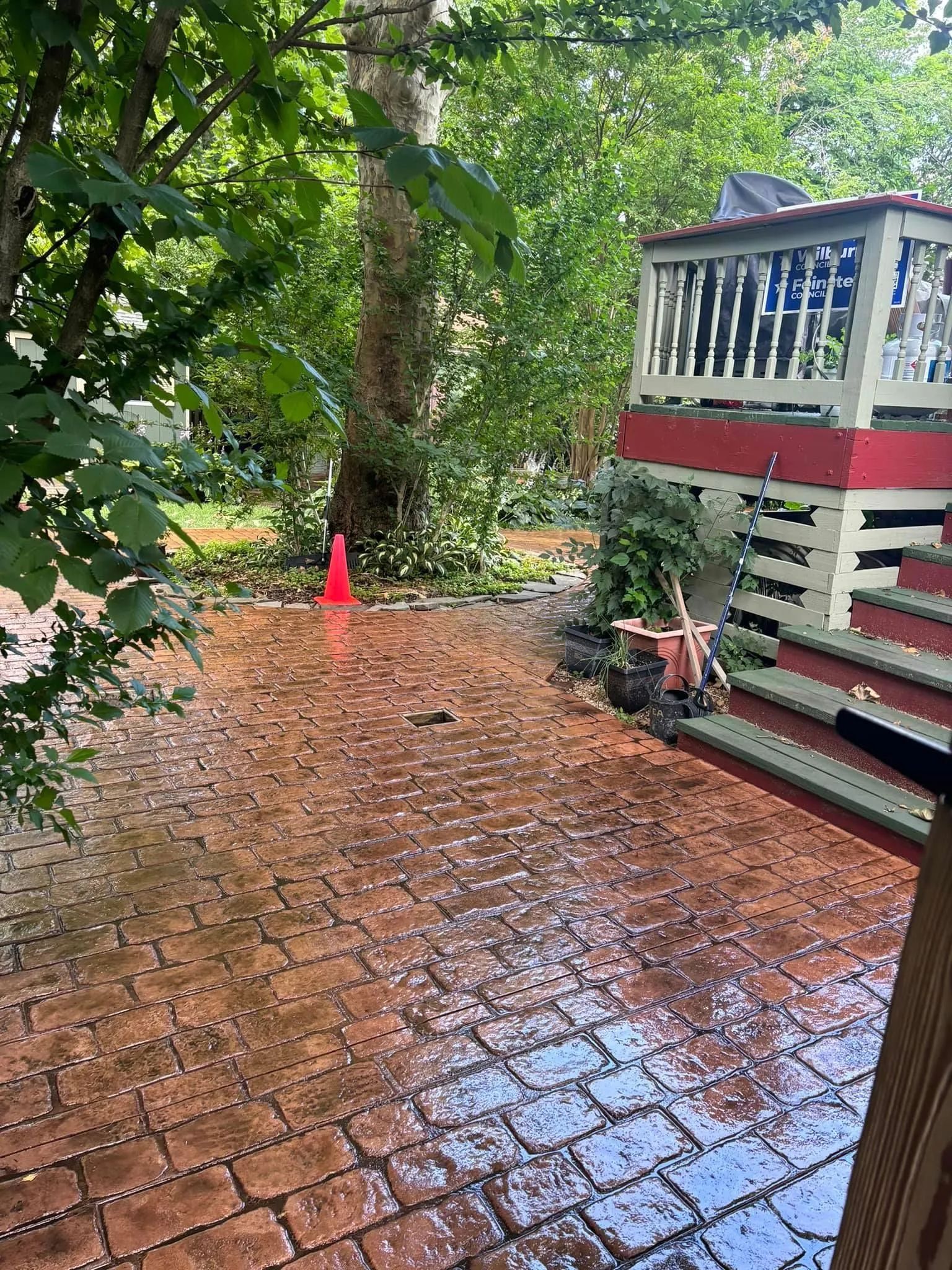A wet brick patio leading toward a raised wooden deck and a large tree in a backyard.