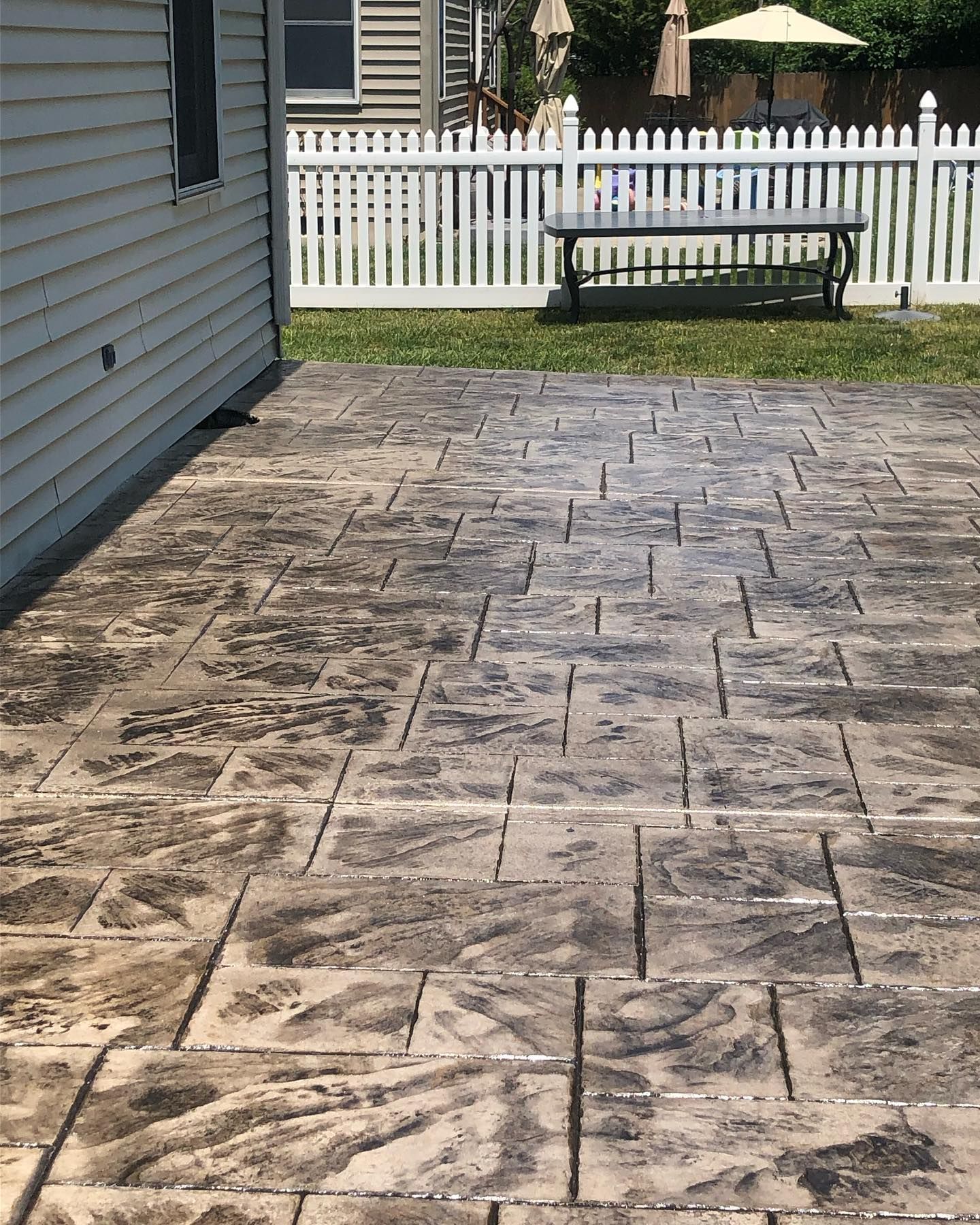 A patio with rectangular stamped concrete tiles next to a house, with a white picket fence and lawn in the background.