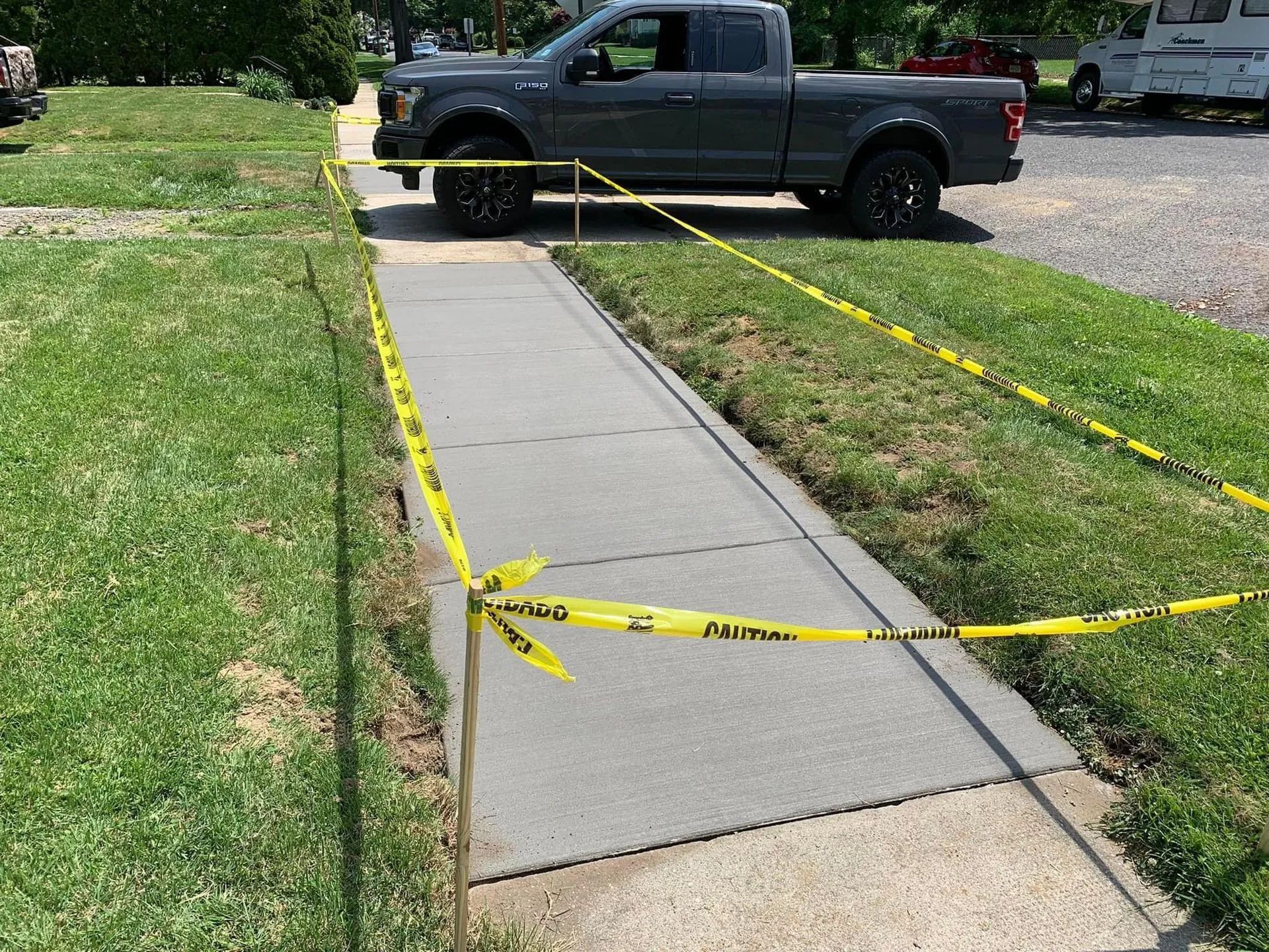 A newly poured concrete sidewalk section is cordoned off with yellow caution tape in a grassy residential area.