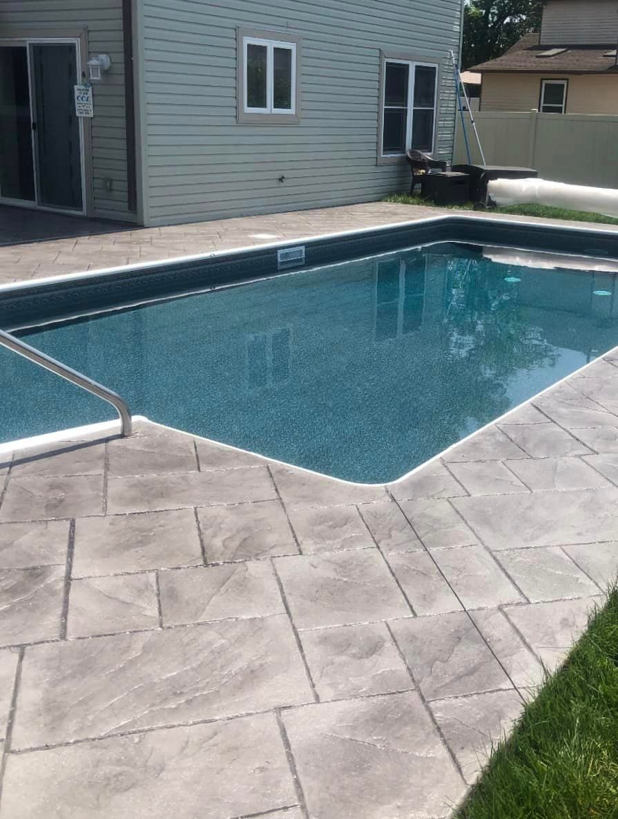 A rectangular swimming pool with blue water and gray patterned concrete decking beside the side of a house.