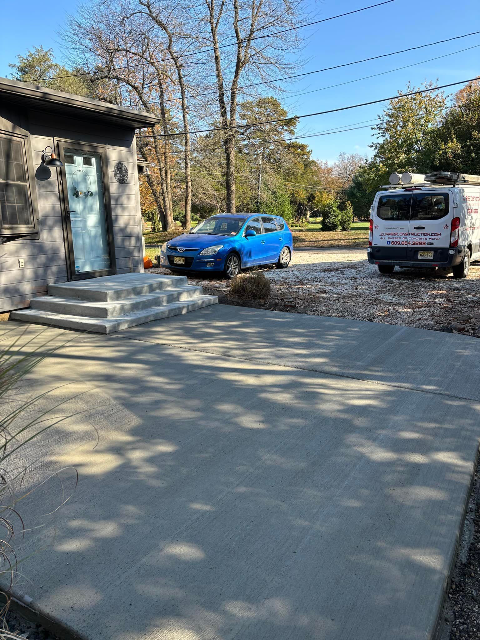 A concrete patio leading to an entryway with stairs, with a blue car and a work van parked on a gravel driveway nearby.