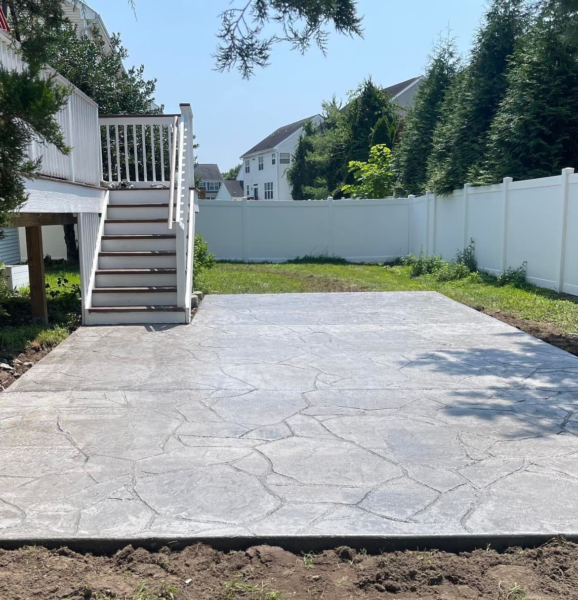 A newly installed gray concrete patio with a stone-patterned design, situated beneath a wooden deck in a grassy backyard.
