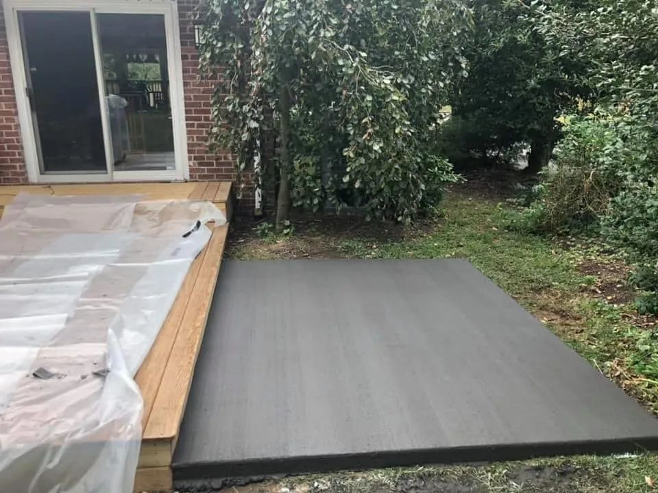 A newly poured gray concrete patio sits beside a wooden deck and brick house wall, adjacent to a grassy, wooded yard.