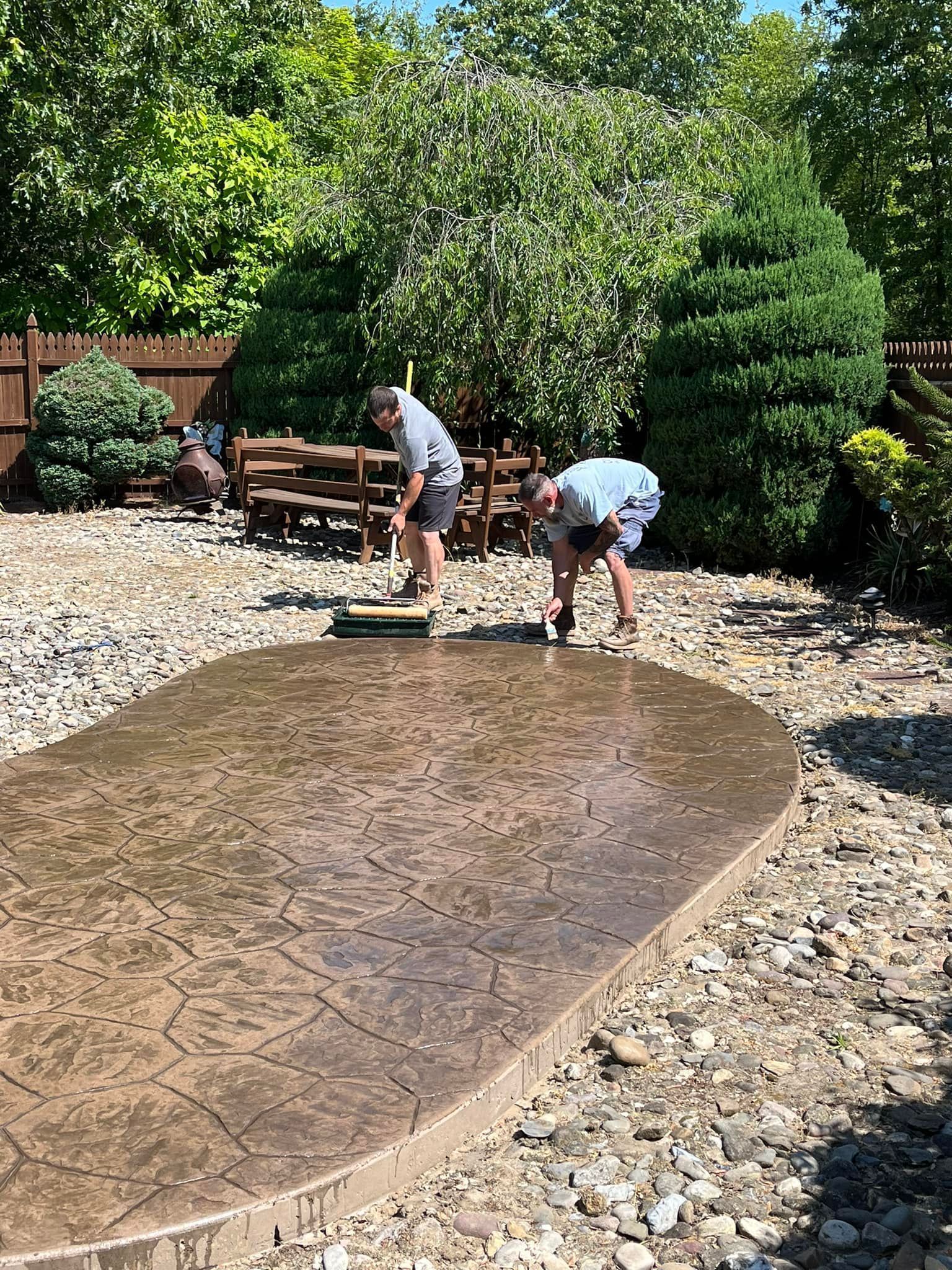 Two people work on a stamped concrete patio in a backyard setting filled with landscaping gravel and greenery.