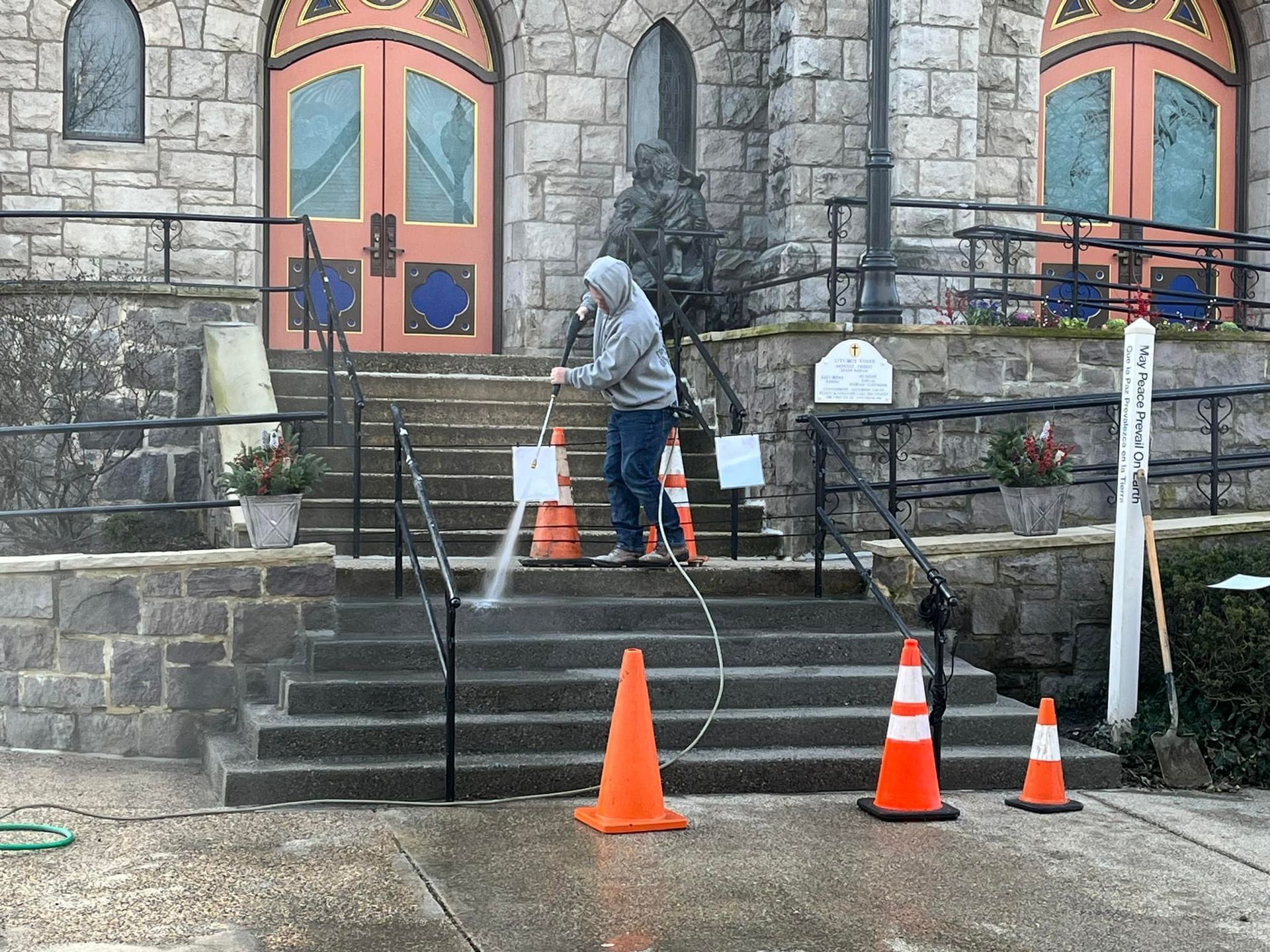A person in a gray hoodie uses a pressure washer to clean the concrete steps of a stone building with orange traffic cones.