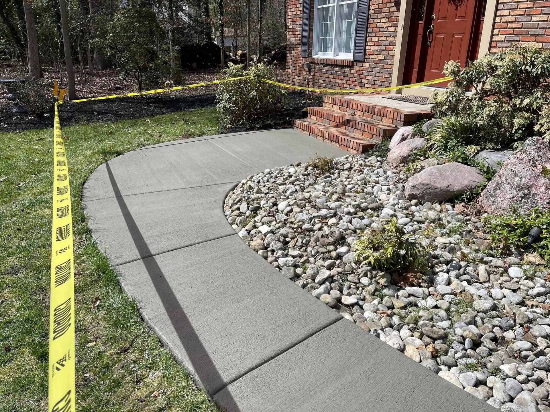A curved concrete walkway leads to brick steps at a house entrance, surrounded by gravel landscaping and caution tape.