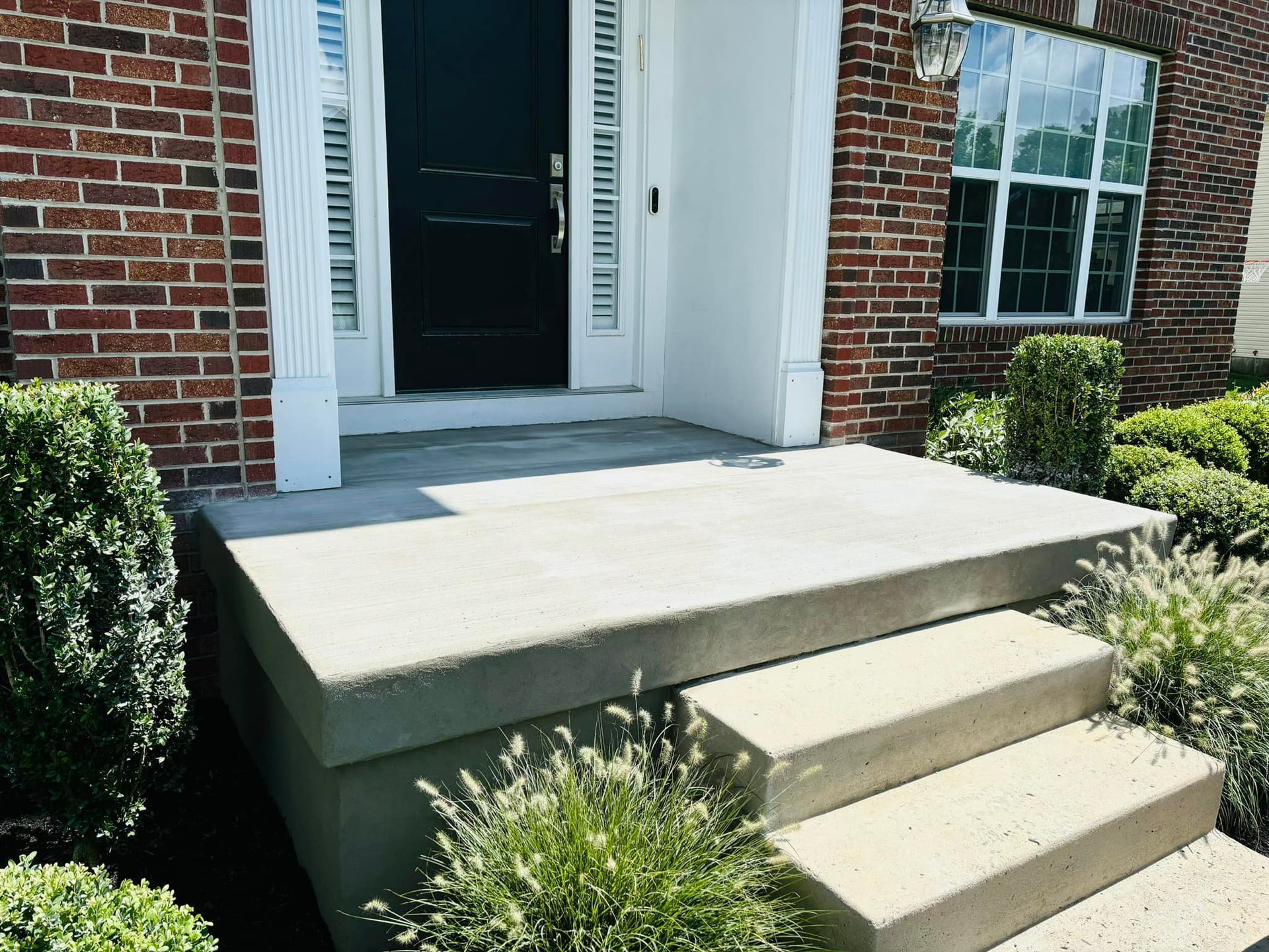 A front entrance with two concrete steps leading to a black door surrounded by white trim and a red brick wall.