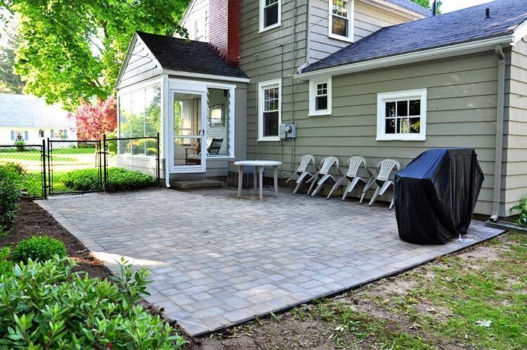 A gray house with a stone paver patio featuring a round table, four chairs, and a covered grill in a grassy backyard.