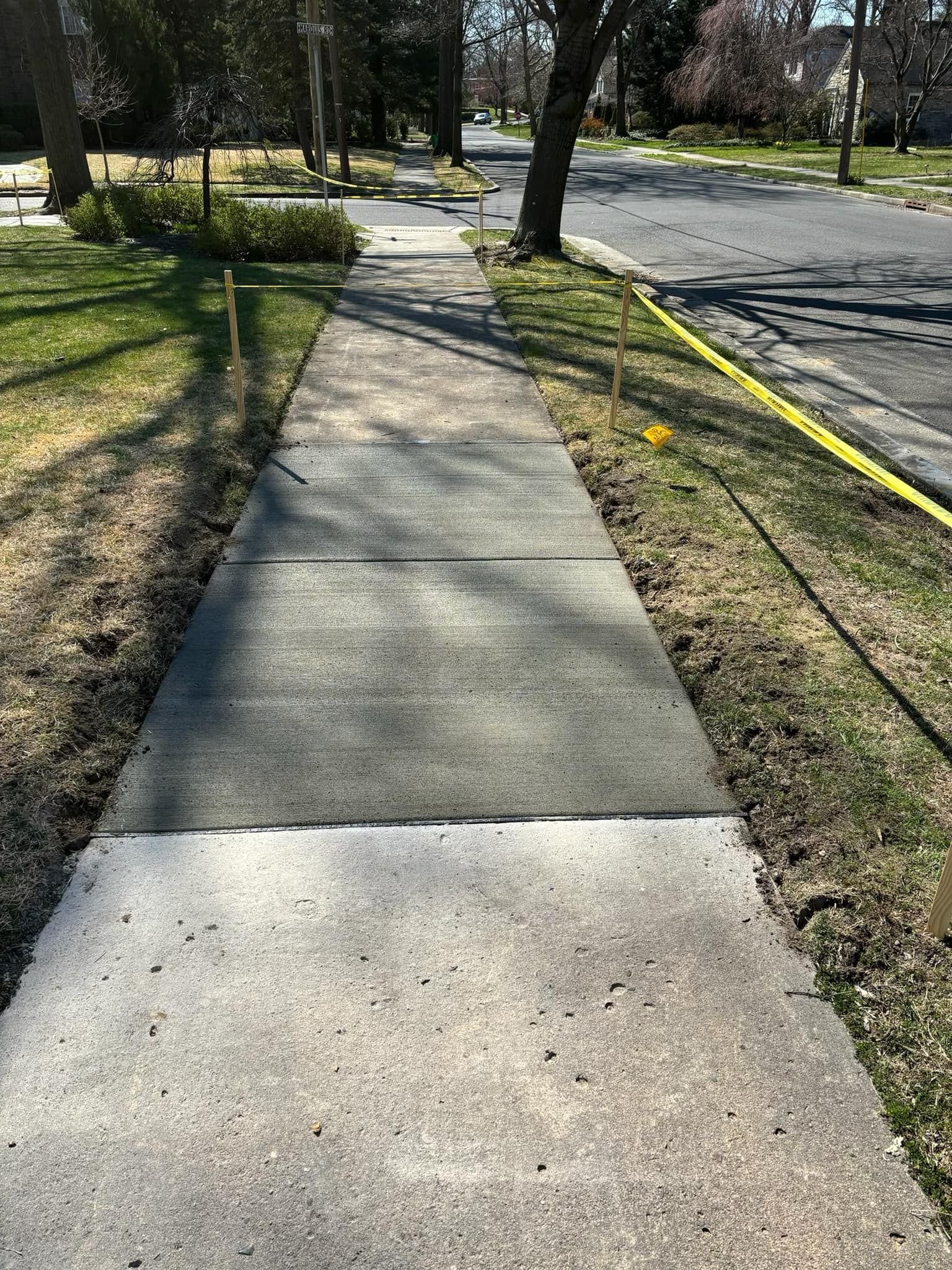 A sidewalk transitions from old, light-colored concrete to a newly poured, darker section marked by thin wooden stakes.