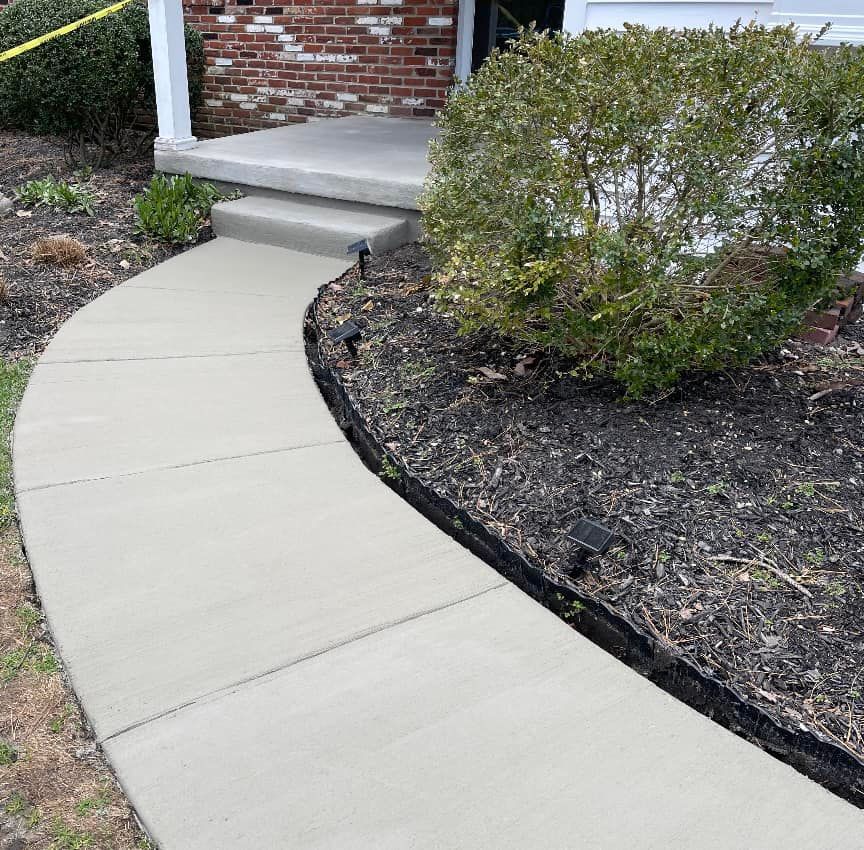 A curved concrete walkway leads to a front porch with brick siding and a large green bush in a mulched garden bed.