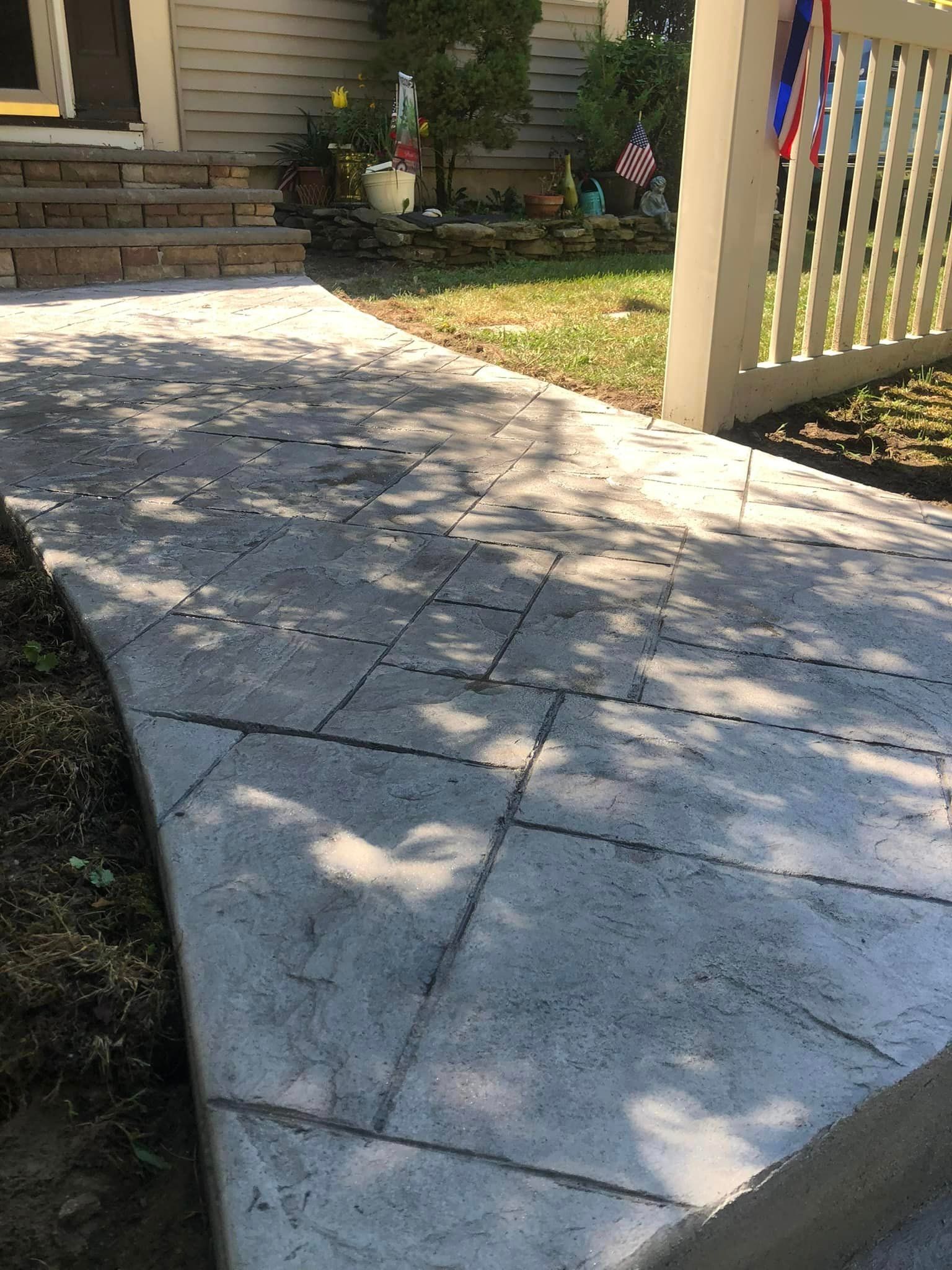 A light gray, textured concrete walkway with a geometric pattern leads toward the front steps of a house.