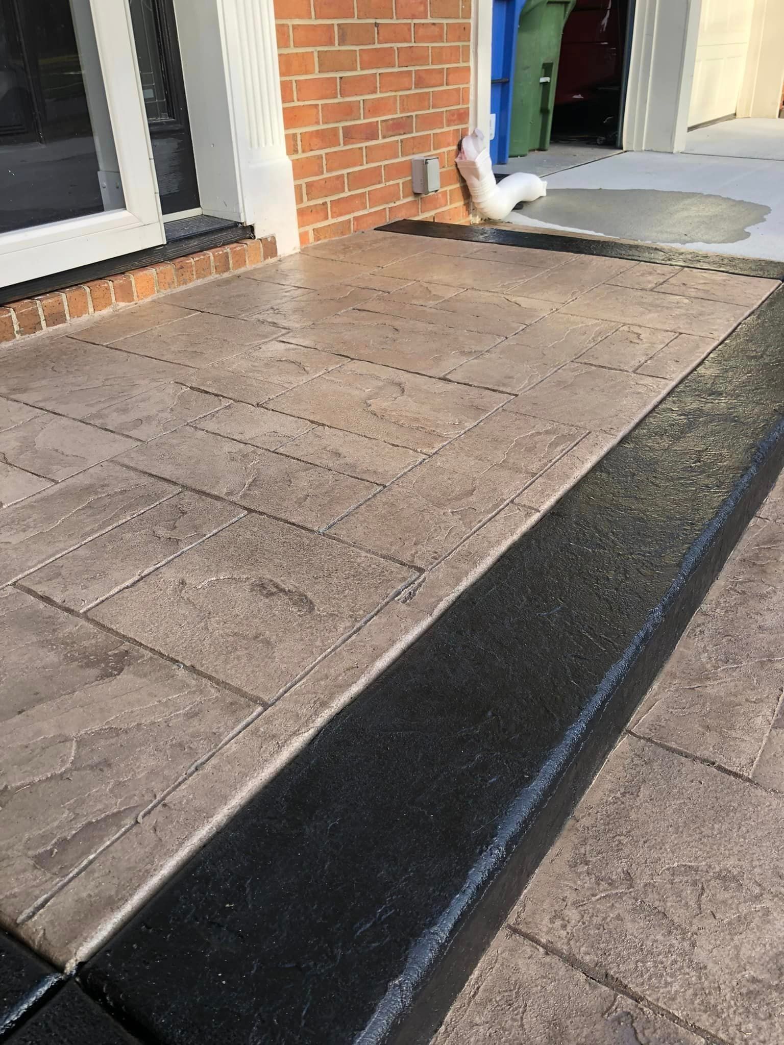 A textured patio with light-brown stone pavers, featuring a dark-gray border and a white downspout extension against brick.