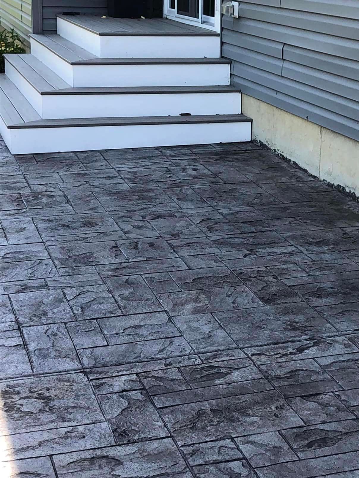 Concrete patio with a stamped stone pattern leading up to three gray wooden steps against a grey-sided house.