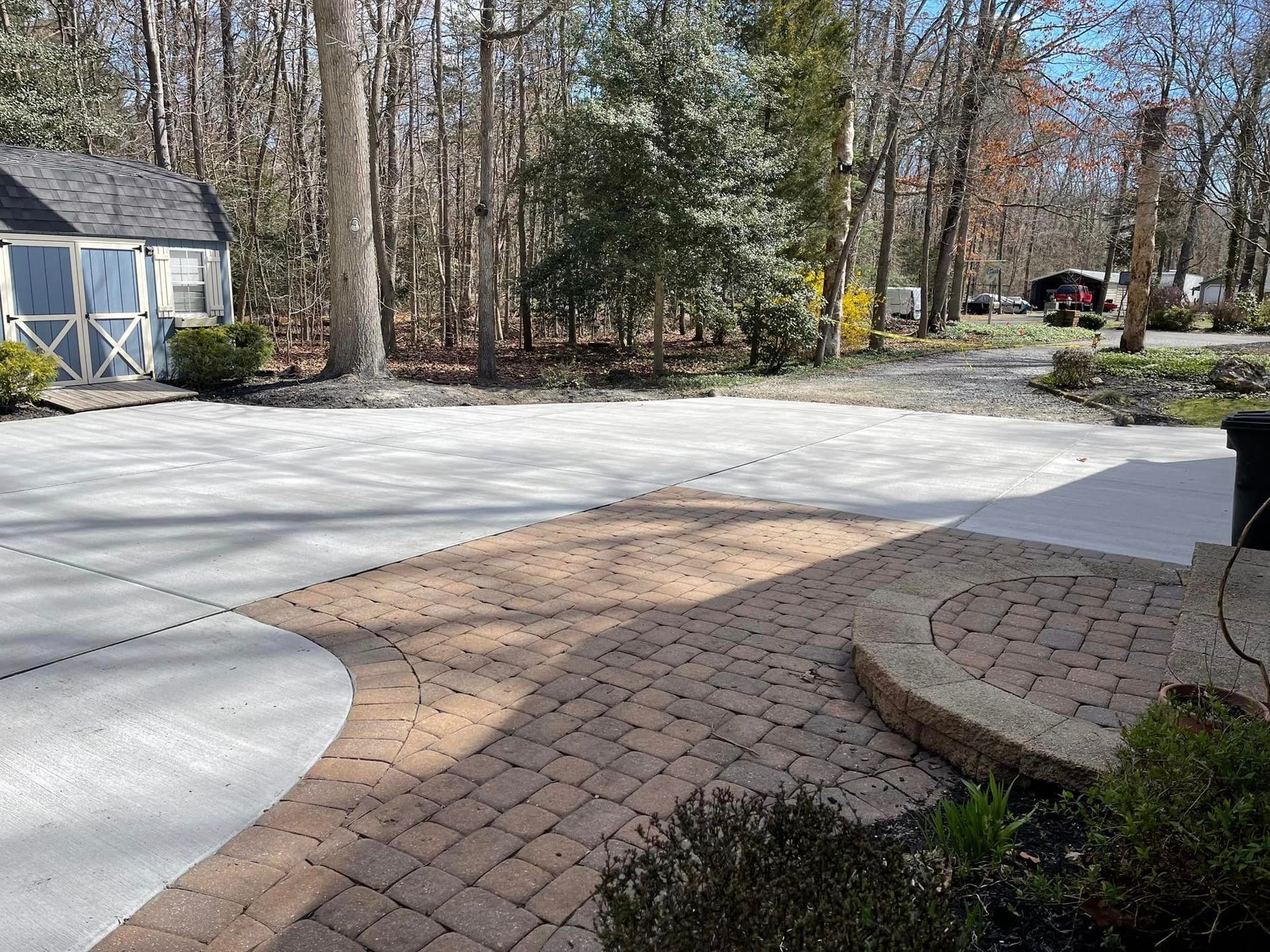 A patio area with brick pavers curving into a concrete driveway, next to a small shed and a wooded backyard.