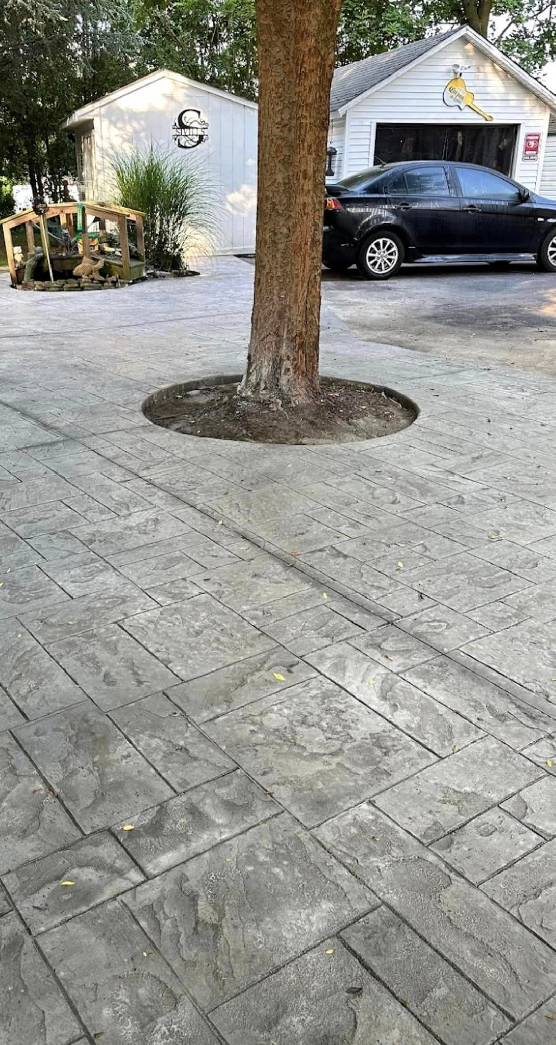 A large tree stands in the center of a gray stamped concrete driveway, with white garages and a parked car in the back.