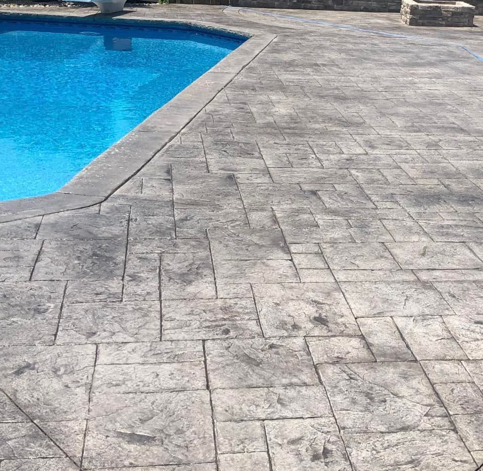 A blue swimming pool edged with gray, rectangular stamped concrete patio pavers in a repeating geometric pattern.