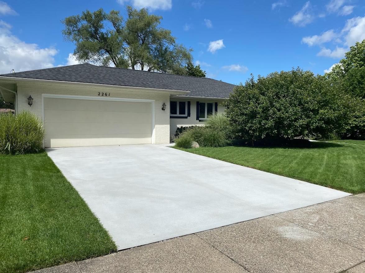 A light-colored house with a dark roof and a freshly paved concrete driveway leading to a closed two-car garage.